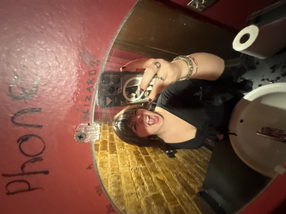 A person takes a selfie in a bathroom mirror. The mirror is round and affixed to a pink wall with black writing. The reflection includes a yellow brick-patterned wall. The person holds a phone with a sticker on its case. They wear rings, bracelets, and a black shirt. The bathroom has a sink, faucet, and a roll of toilet paper.