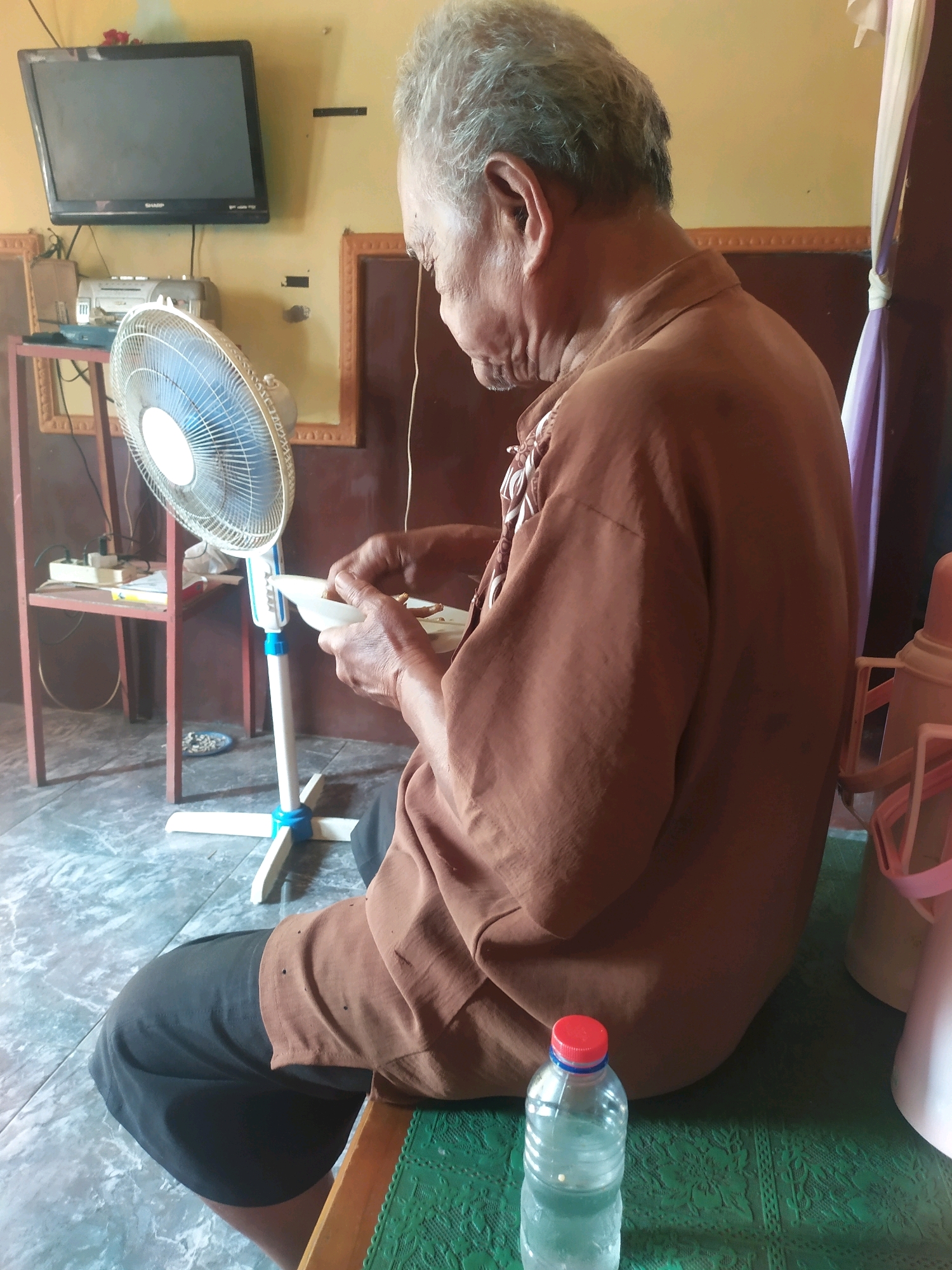 An elderly man with gray hair sits on a green mat in a room that appears to be in a home in Desa Penggalang, Kecamatan Adipala, Indonesia. He is wearing a brown collared shirt and dark pants. The man is holding a white bowl in his left hand and appears to be eating from it. In front of him is a white standing fan with a blue base. To his left, on a shelf, is a black television set and a radio. A plastic bottle of water with a red cap is visible in the foreground. The room has beige walls and tiled flooring. The overall impression is that of a simple, domestic setting.