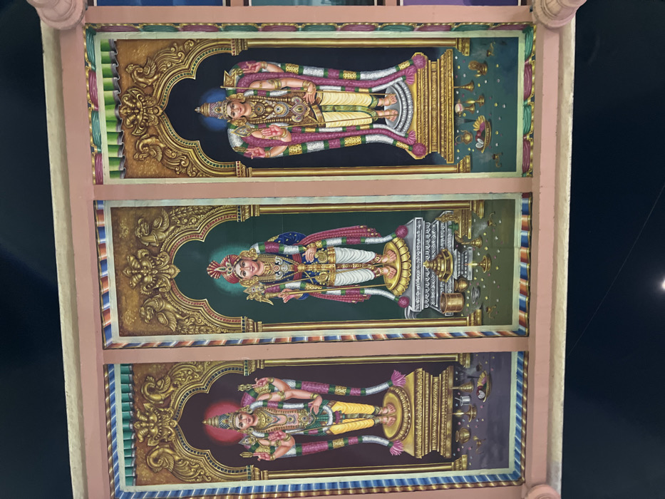 The image displays a vibrantly painted ceiling inside what appears to be a Hindu temple in Palani, India. Three rectangular panels, aligned vertically, each feature a standing deity rendered in a classical South Indian artistic style.
The uppermost panel showcases a four-armed deity, possibly Vishnu or a similar form, with light blue-green skin. This deity wears a tall golden crown, intricate jewelry, and draped garments, holding traditional attributes such as a conch, discus, and mace. He stands on a golden pedestal against a light green background, flanked by depictions of ritual items.
The central panel depicts a two-armed deity, identifiable as Murugan (Kartikeya or Skanda), a prominent deity in South India. He is adorned with a distinctive turban-like crown, elaborate ornaments, and holds his characteristic weapon, the Vel (spear). His skin is fair, and he stands on a pedestal against a darker green backdrop, with offerings arranged beside him.
The lowermost panel presents another four-armed deity, resembling Vishnu, with fair skin and a prominent red halo behind his head. He, too, is richly attired with a crown, jewelry, and garments, holding various divine attributes. This deity stands on a pedestal against a dark purple-blue background, accompanied by ritualistic offerings.
Each deity painting is framed by an ornate, gilded archway featuring elaborate scrollwork and decorative motifs, emphasizing the grandeur. The panels are set into a larger structural frame, bordered by colorful bands of pink, blue, and green, suggesting the rich architectural and artistic traditions of a temple interior. The deities are depicted with serene expressions, evoking a sense of calm and devotion. No text is visible in the image.