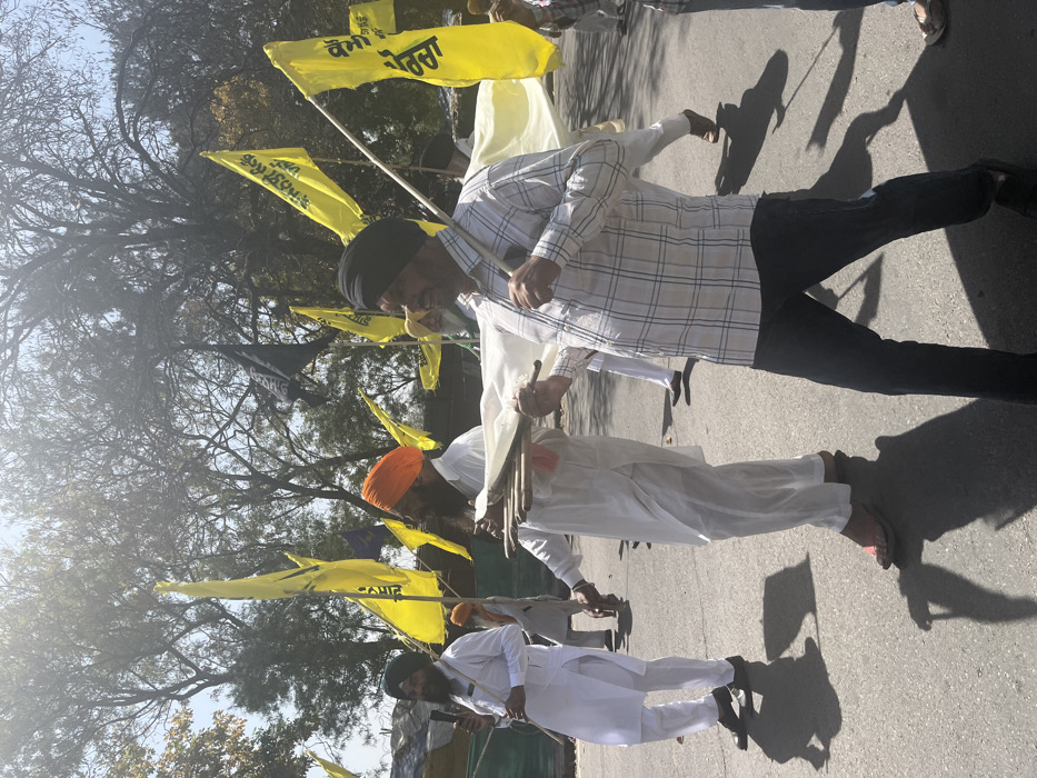The image shows a group of men walking down a paved street in Sahibzada Ajit Singh Nagar, India. The men are wearing white kurtas, turbans, and black pants. They are holding wooden sticks and a white cloth banner, which is being carried by a man in a plaid shirt, who is at the forefront of the group. The man is walking towards the camera with a confident stride. There are several yellow flags with Indian writing visible in the background. The sky is blue with white clouds, and there is a tree in the distance. The men seem to be participating in a procession.