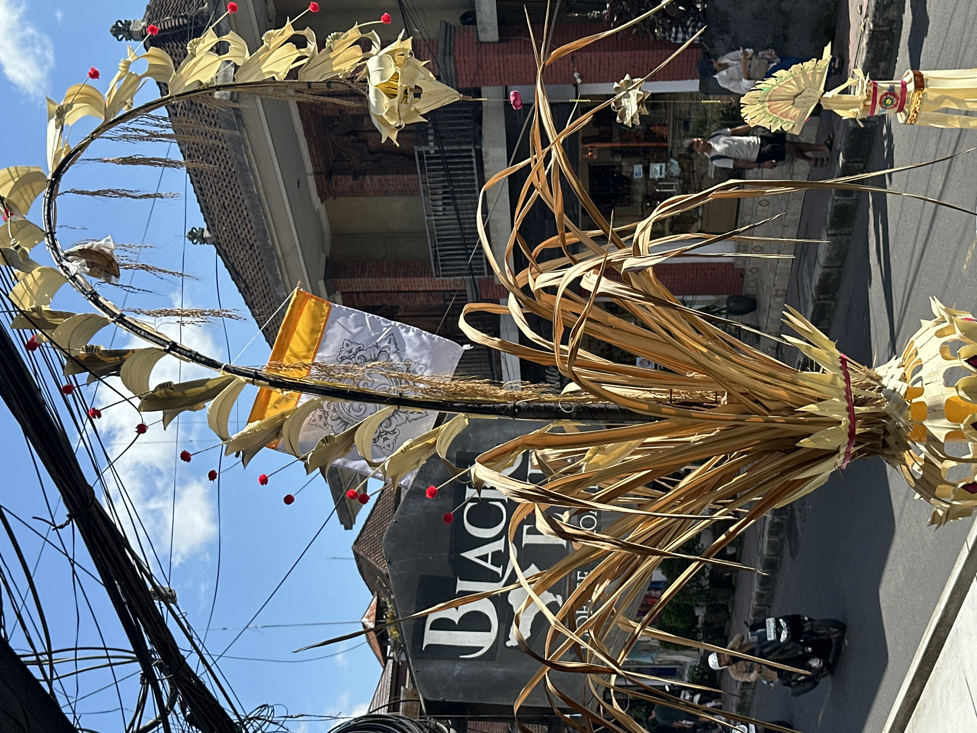 This is a full, eye-level, vertical shot of a street scene in the Ubud District, Indonesia, on a sunny day. The foreground is dominated by a large, dried palm frond decoration with a yellow and white flag attached, which is arched and adorned with small red pom-poms. Behind this decoration, a street runs horizontally, with a sidewalk on the right. A black sign with the white lettering "BLACK TEA" is visible on a building across the street. Several people are present in the scene: a person on a scooter is riding down the street, and two people are walking on the sidewalk. Electrical wires are strung across the top of the image, creating a cluttered overhead. The buildings in the background have traditional Balinese architecture.