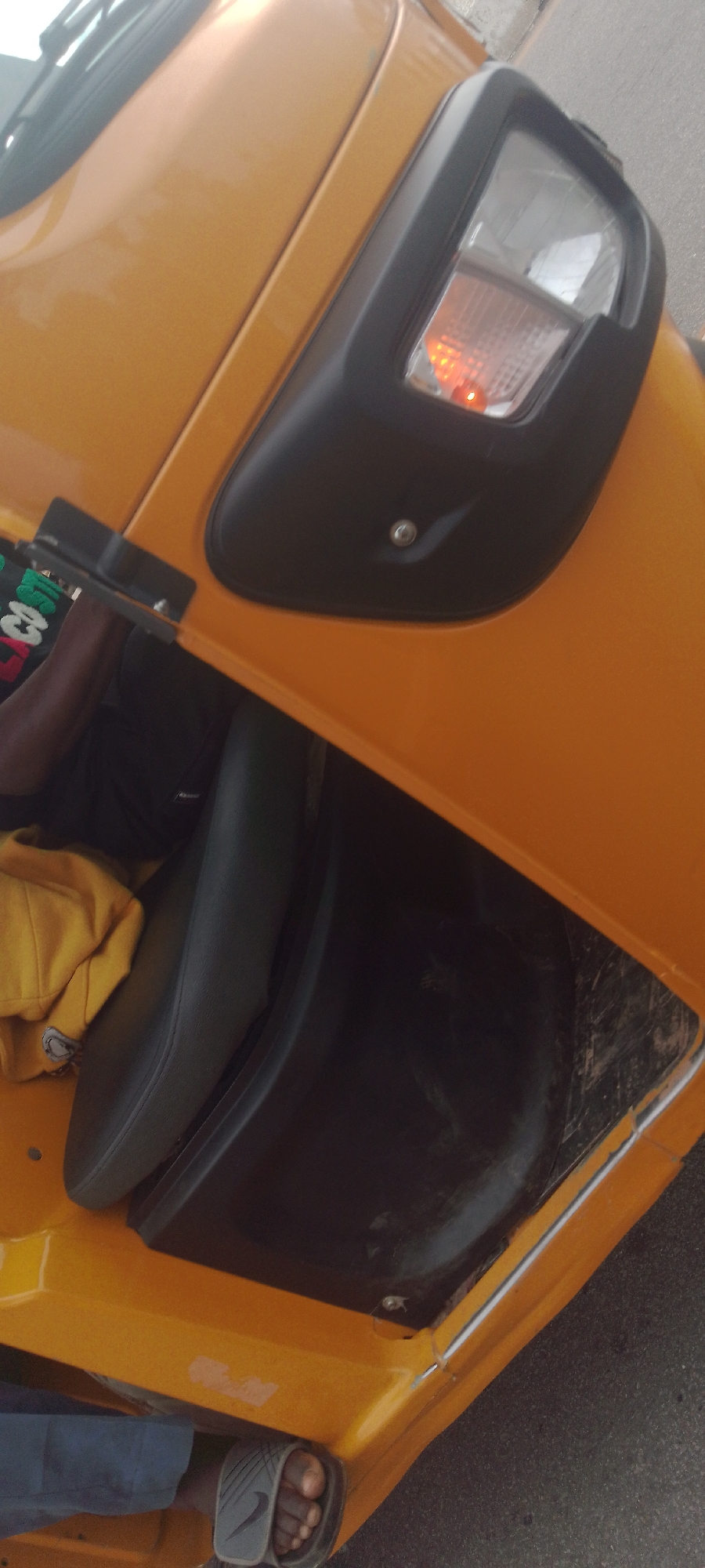 A tilted, close-up shot focuses on the bright orange exterior of a three-wheeled vehicle, likely an auto-rickshaw or "Keke Napep," in Jalingo, Nigeria. The vehicle's bodywork is prominently featured, with a black plastic housing enclosing the headlight and a small orange indicator light. Visible within the vehicle's open side is a dark-colored seat, partially covered by a folded yellow garment. A person's arm and shoulder are visible near the top left, suggesting they are operating or interacting with the vehicle. A person's foot, wearing a gray flip-flop with a Nike logo, is visible at the bottom left, resting on the vehicle's orange floorboard. The background is out of focus, showing a glimpse of a paved surface, likely a road, and indistinct elements suggesting an outdoor, urban setting. The overall impression is of a functional, everyday vehicle in use within its likely environment.