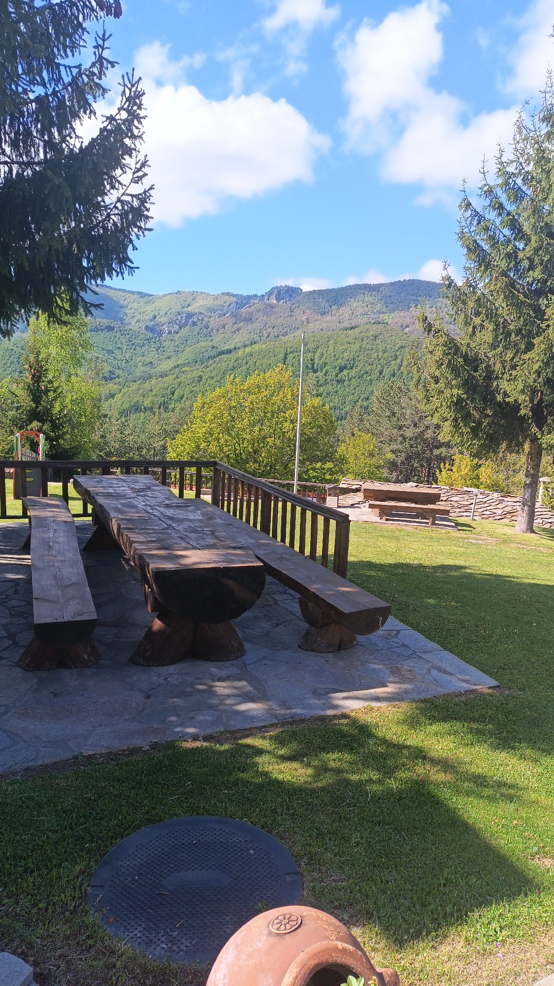 The image depicts a large, wooden picnic table with benches on a stone patio. The table and benches appear to be constructed from massive logs. A wooden fence surrounds the patio area, offering a view of a mountainous landscape covered with trees, under a blue sky with scattered clouds. In the foreground, near the bottom of the image, there's a dark, circular object, possibly a manhole cover, and part of a terracotta pot. The location is Vrata, Bulgaria, suggested by the mountainous landscape consistent with the area. There are no people visible in the image. The scene is outdoors, suggesting a recreational or dining area in a scenic location. Another table and bench set can be seen on the grass near a low rock wall.