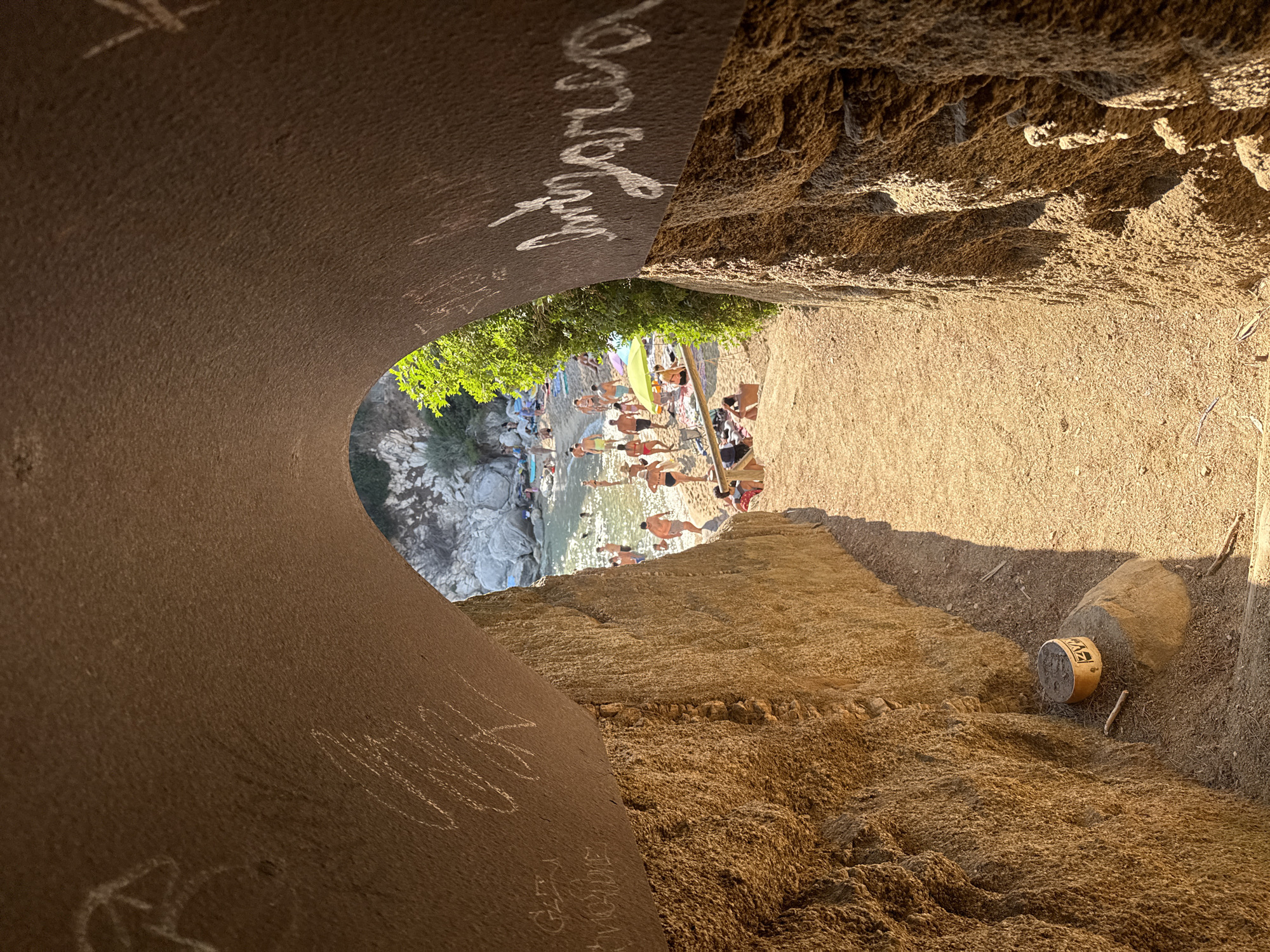 This image captures a view from within a cave or rock formation looking out onto a crowded beach. The foreground is dominated by the textured, rough walls of the cave, which are inscribed with white chalk markings. The cave opening offers a framed perspective of the bustling beach scene beyond.
On the beach, numerous people are visible, dressed in swimwear. Some are standing in the shallow, clear water, while others are on the sand. Beach umbrellas and towels are scattered across the sand, indicating a popular day for sunbathing and swimming. The water appears calm and inviting, reflecting the sunlight. The background consists of rocky outcrops and some green foliage, typical of a Mediterranean coastline. The overall atmosphere suggests a vibrant and active beach environment.