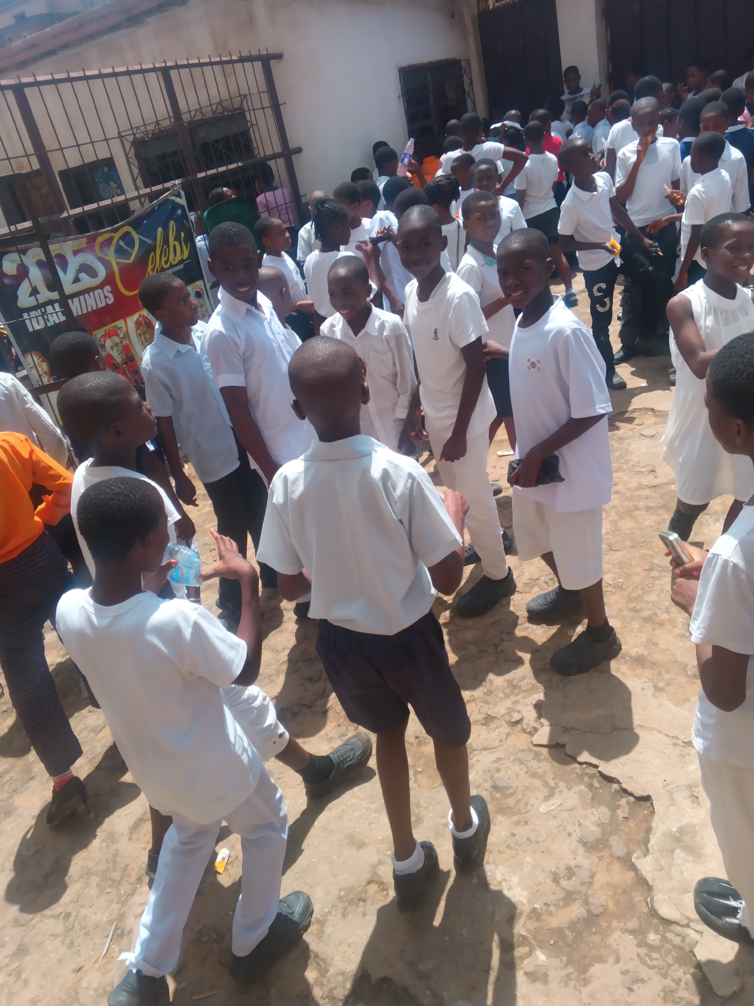 This media file depicts a gathering of approximately thirty children, mostly boys, in an outdoor setting. The children are predominantly dressed in white attire, some in shirts and trousers, and others in shorts. A few are wearing white shirts with what appear to be school uniforms, indicated by small badges or insignias on their chests, one of which resembles the South Korean flag.
The scene appears to be a public space, possibly outside a building with a barred entrance. A large banner with the text "2025 CELEBI" and "IDEAL MINDS" is visible, suggesting an event or campaign related to education or youth development. The ground is uneven and dusty, with patches of dry earth and some debris. The lighting suggests it is daytime, with strong sunlight casting shadows, implying a sunny weather.
Some children are interacting with each other, while others are looking towards the camera or elsewhere. One boy in the foreground is holding a bottle of water. Another child is holding a mobile phone. The overall mood seems to be one of excitement or anticipation, as suggested by the energetic poses and expressions of some of the children. The presence of a banner and a large group of children in uniform implies a school-related event, possibly a celebration, competition, or gathering. The location context provided places this scene in Nkpor, Nigeria.