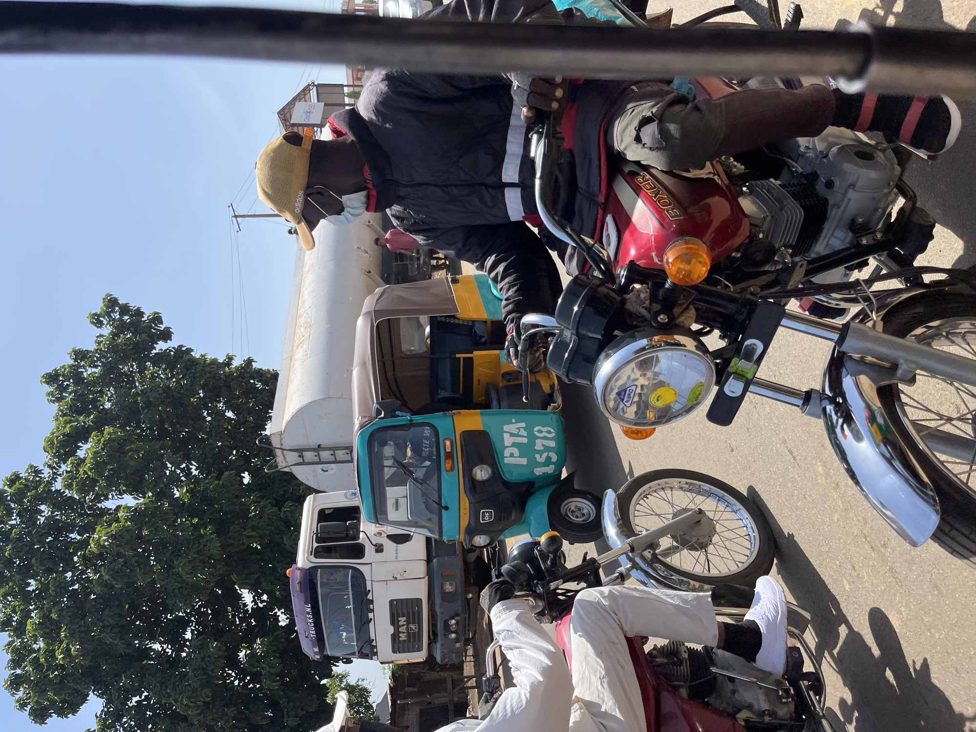 This image captures a bustling street scene in Jos, Nigeria, during bright daylight under a clear blue sky. The perspective is low, looking forward from what appears to be another vehicle.
In the immediate foreground, a person is seated on a red "BOXER" brand motorcycle. This individual, likely male, is wearing a black puffer jacket, a light-colored Adidas baseball cap, and a light blue surgical mask. Black gloves with white stripes cover their hands, and their left leg shows dark, possibly ripped, pants. They wear black and red striped socks with dark shoes. To the bottom right, the legs and feet of another person, wearing white pants and white sneakers, are visible, seated on a similar red motorcycle.
The middle ground features a turquoise and yellow motorized tricycle, commonly known as a Keke Napep, with "PTA-1578" clearly visible on its side. Behind the tricycle is a large white tanker truck, bearing the "MAN" logo on its cab and "WWW.TRUCKS.NL" printed below its windshield. Another truck is partially visible further behind the tanker.
The scene unfolds on a paved road, with strong shadows cast by the vehicles, indicating sunny weather. A large, verdant tree occupies the left side of the frame, contrasting with the urban environment. In the distant background, a billboard with "LED" visible can be seen. The overall impression is of a busy street with vehicles possibly stopped in traffic.