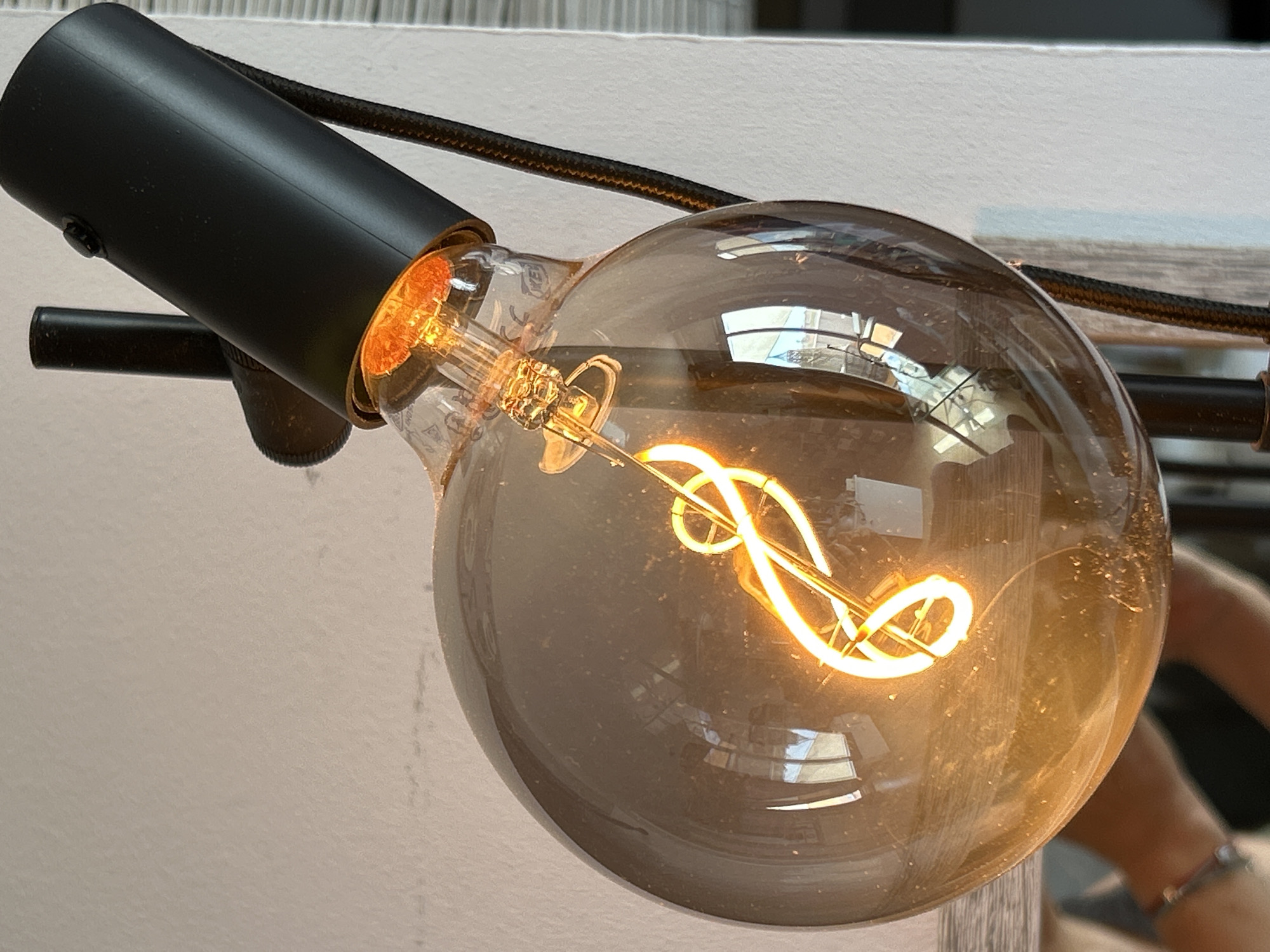 The image shows a close-up view of a vintage-style Edison light bulb, glowing warmly. The bulb has a visible filament coiled in a spiral pattern. It is housed in a black, angled lamp fixture, which appears to be connected to a black, fabric-covered cord. The background consists of a white wall and a textured surface, possibly a vertical blind or wood paneling. Reflections of a room, including what looks like windows and furniture, are visible on the surface of the glass bulb. A person's arm, wearing a bracelet, is partially visible in the lower left corner of the frame, suggesting the lamp is part of an indoor setting. The image does not provide any specific information or context related to the city of Pilar de la Horadada, Spain.