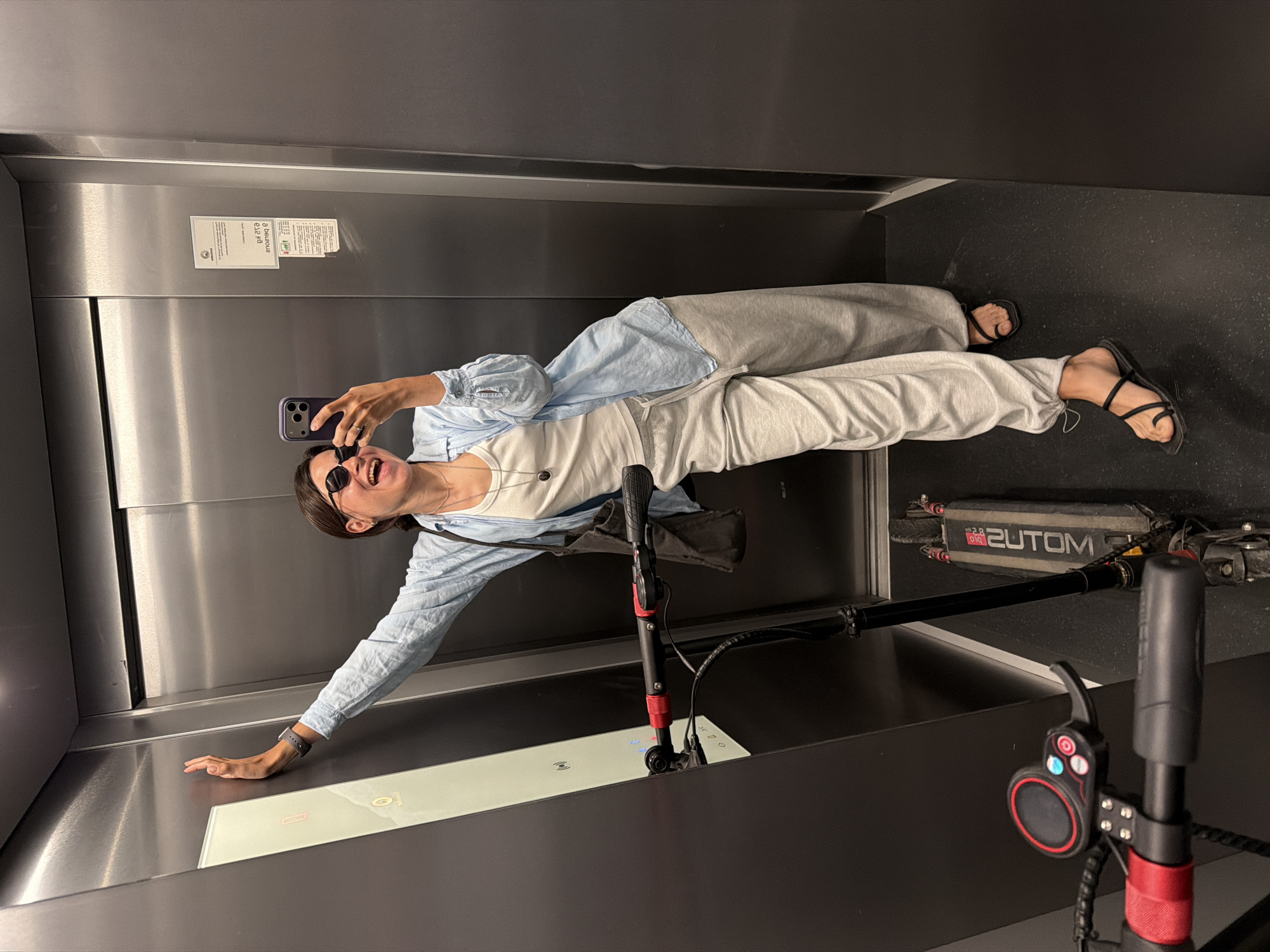 A person lies in an elevator taking a selfie with their smartphone. They are wearing sunglasses, a white shirt, light blue button-down shirt, and light gray sweatpants. The person has dark hair pulled back. Next to them is a black electric scooter, with the brand name "2UTOM" visible. The elevator has stainless steel walls and a black floor. A control panel with buttons is visible. There's a sign on the wall. The city of Budva, Montenegro is not discernible from the image.