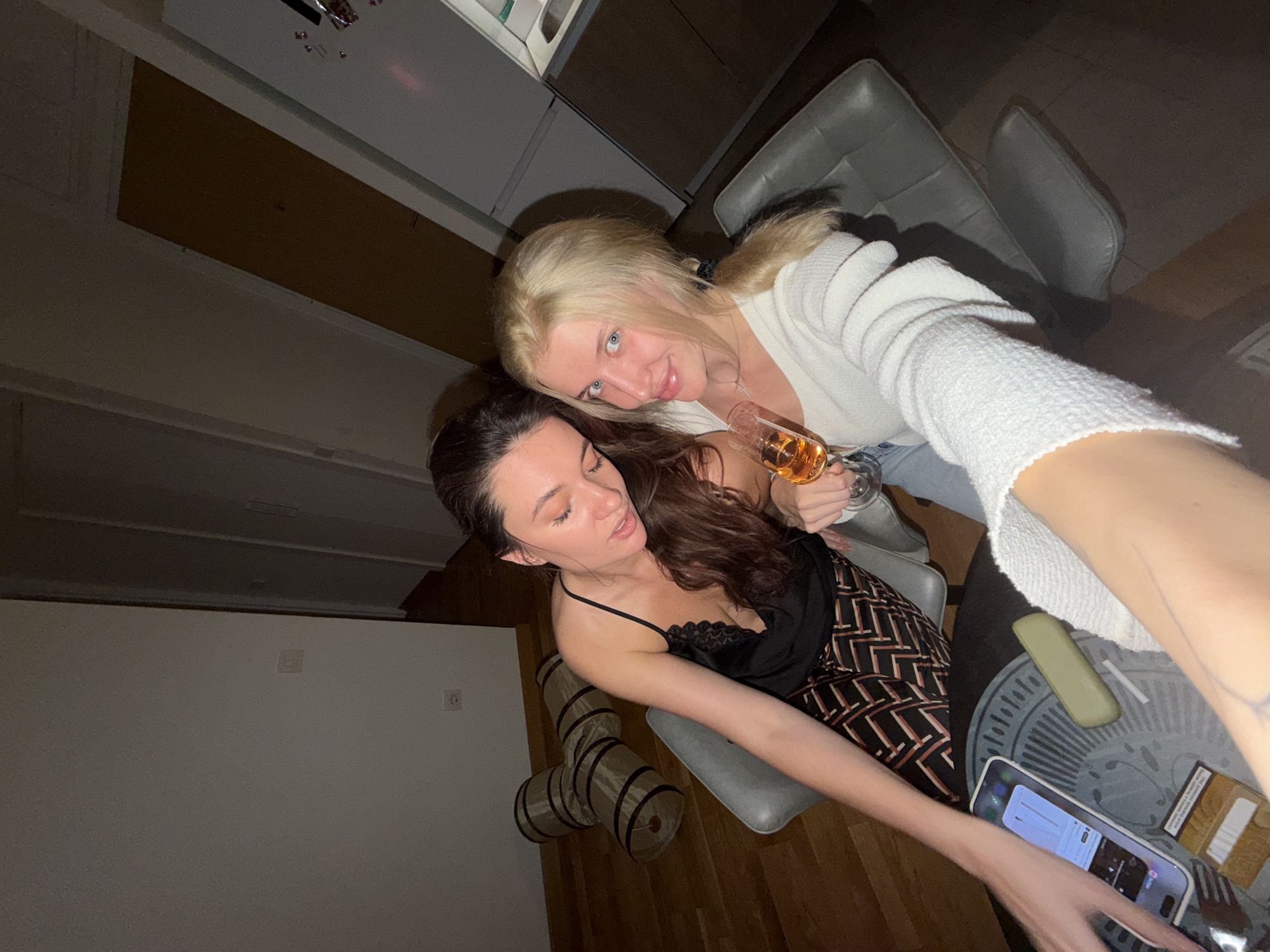 Two women pose for a selfie. One woman, with long blonde hair, wears a white sweater and holds a wine glass. The other woman, with long brown hair, wears a black top with lace and patterned pants. They are seated. A table with a phone, a cigarette pack, and a lighter is next to them. A gray chair is in the background. There's also a cat tunnel toy in the background, near a blank white wall. The location is unknown.