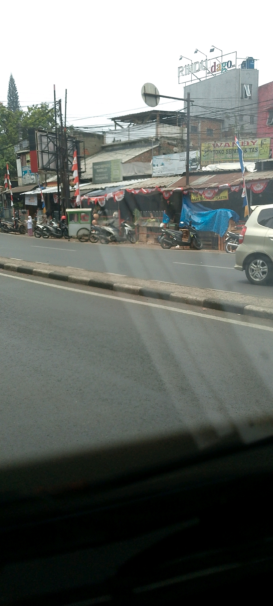 The image captures a street scene in Bandung City, Indonesia, likely on a main road with shops and traffic. In the foreground, a dark tinted windshield creates a silhouette, obscuring the lower portion of the scene. Beyond this, a road surface with a light-colored curb and white markings is visible, indicating a traffic lane.
The middle ground reveals a row of low-rise buildings and shops, many with corrugated metal roofs. There are several motorcycles parked along the roadside and a few people are visible. One individual in a white hijab stands near a green and white food stall. Another person on a red motorcycle is visible further back, and several other motorcycles are parked or in motion. A white car is parked on the right side of the road, facing away from the viewer. Some of the shops have awnings and decorations, including red and white banners that suggest a national celebration or event. Signs are visible above the shops, with one prominent sign on a grey building reading "Rindu dago" in colorful lettering, indicating a commercial area.
The background shows more buildings and structures, along with overhead power lines and a tall, slender evergreen tree on the left. The sky is overcast, suggesting a cloudy day. The overall impression is of a bustling urban environment with typical street commerce and traffic.