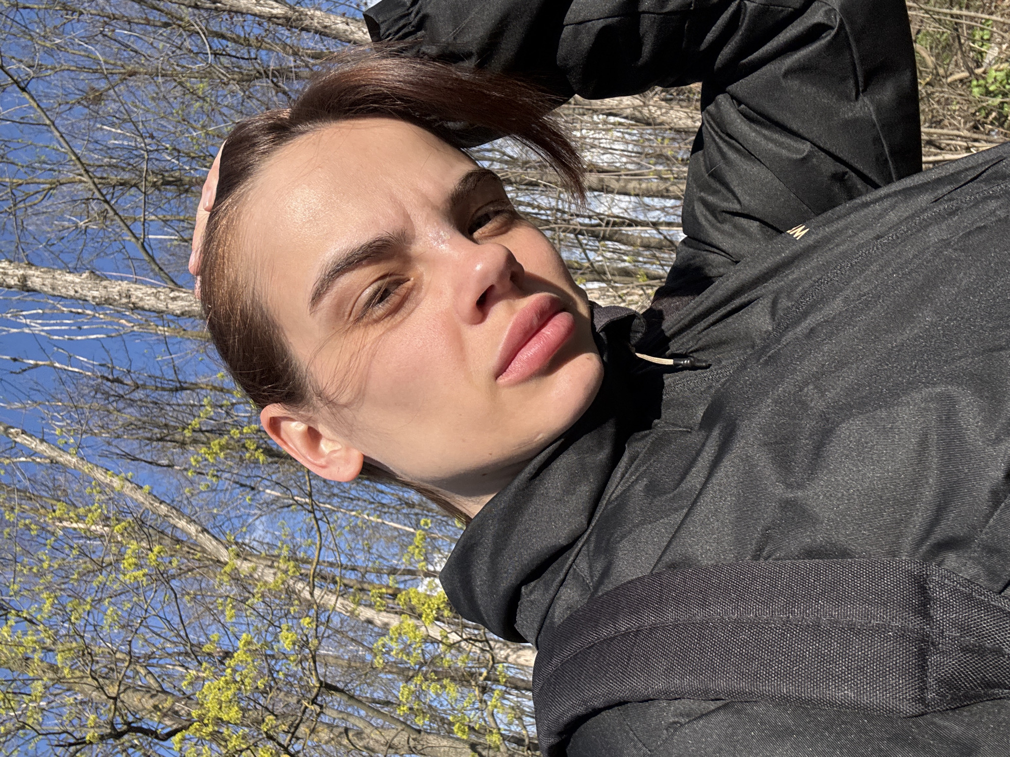 A young woman stands outside on a sunny day with trees in the background and blue sky visible through the branches. The woman is fair-skinned, with brown hair. She wears a black puffy jacket. She is holding her hand on her forehead. The location is likely outdoors in an area with trees and foliage, but specific details about Rostov-on-Don cannot be determined from the image.