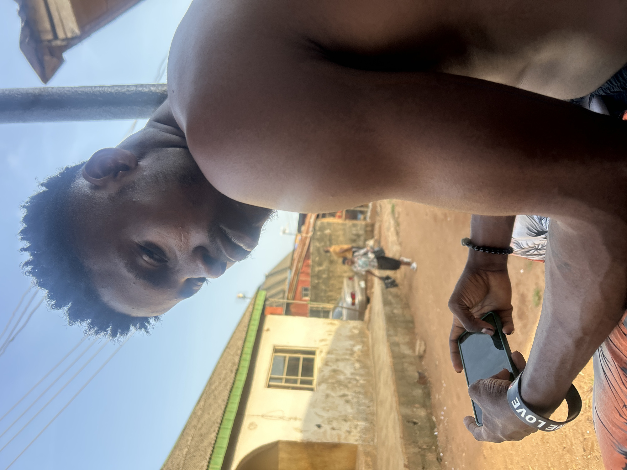 A brown-skinned man is holding a smartphone and looking at the camera. He has short black hair and is wearing a black rubber bracelet that says "LOVE". The background shows a street scene with a building on the left, and a figure walking away in the distance. The location appears to be an urban area, though the city is unspecified. The scene is bathed in daylight.