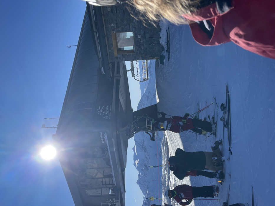The image captures a vibrant, sunny day at a ski resort, rotated 90 degrees clockwise from its original orientation. The perspective is first-person, looking down past a red jacket with a fur-lined hood.
The primary subject is a modern ski lift station, dark grey or black, with a stone-clad building section. Visible text on the station clearly reads "La Rosière" accompanied by a sun logo, "Espace San Bernardo," and "MONT VALAIS." The sun is prominently featured on the left, emitting a strong glare and lens flare against a clear, bright blue sky.
In the foreground and mid-ground, several individuals are visible in full ski and snowboard attire. Many wear red jackets, while one person is in a dark jacket and tan pants, equipped with a snowboard. Others are on skis, holding ski poles. They appear to be queuing for or standing near the ski lift, ready to ascend the mountain.
The setting is a vast snow-covered mountain landscape, with distant peaks visible under the brilliant blue sky. The bright conditions suggest it is midday, perfect for winter sports. The scene conveys an active and picturesque environment typical of a high-altitude ski destination like La Rosière in Séez, France, which is part of the Espace San Bernardo ski area.