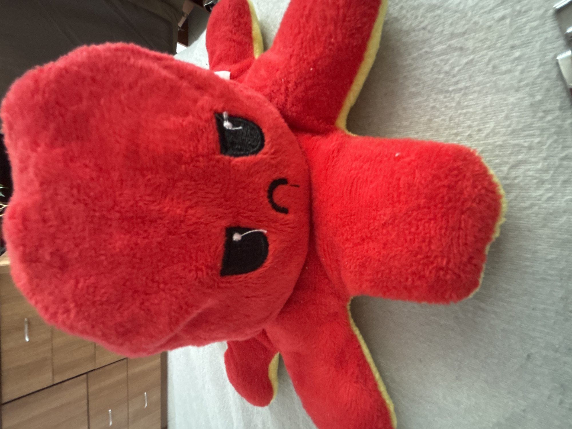 A close-up shot of a red, plush octopus toy with a yellow border on a gray surface. The octopus has black, cartoonish eyes and a simple curved mouth. The background includes a light-colored wooden cabinet and a glimpse of what appears to be a bed. The location in Troyan, Bulgaria, cannot be determined from the visual information.