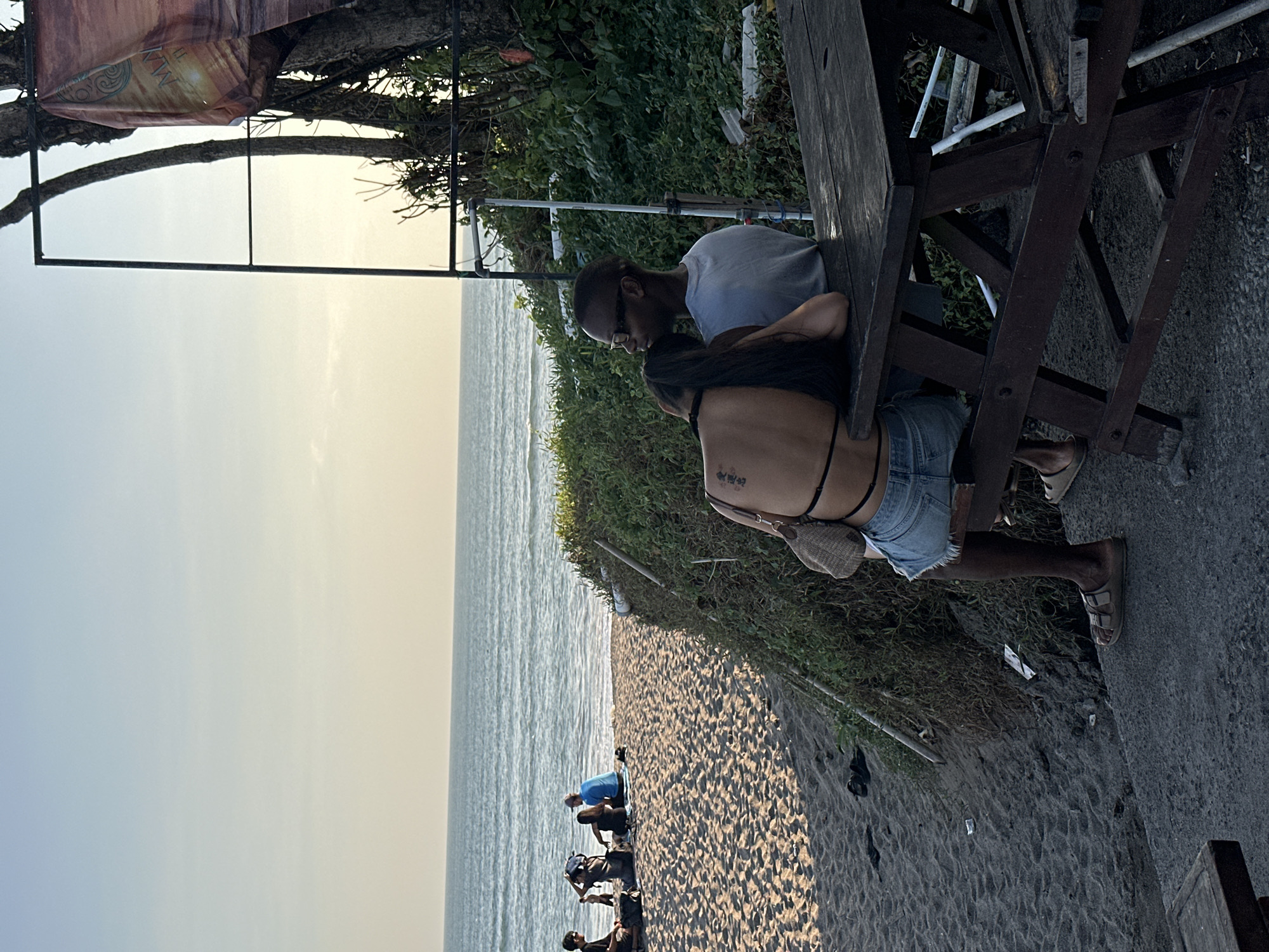 In this image, a group of people are gathered on a beach in North Kuta, Indonesia, near a wooden picnic table. The sun is low in the sky, suggesting it is either early morning or late afternoon, casting a warm, soft light across the scene. The sky is a gradient of pale blue and yellow. The ocean, with gentle waves, stretches out to the horizon, and the beach is covered in dark sand. Lush green vegetation grows behind the picnic area, adding a vibrant contrast to the sand and sea.
Two individuals are prominently featured at the picnic table. A man with glasses and a light-colored t-shirt is leaning over the table, looking down. Beside him, a woman with long dark hair is seated, wearing denim shorts and a dark top. Her back is to the camera, and she appears to be interacting with the man.
Further down the beach, several other people are visible. Some are sitting or kneeling on the sand, while others are standing. One person in a blue t-shirt is kneeling on the sand, seemingly engaged in an activity with others around them. The overall atmosphere appears relaxed and casual, typical of a beach setting.
A banner is visible hanging from a tree or structure in the upper left corner of the image. The banner has an abstract design with warm colors, and some text is present, but it is partially obscured and difficult to read clearly. The scene conveys a sense of leisure and enjoyment in a tropical coastal environment.