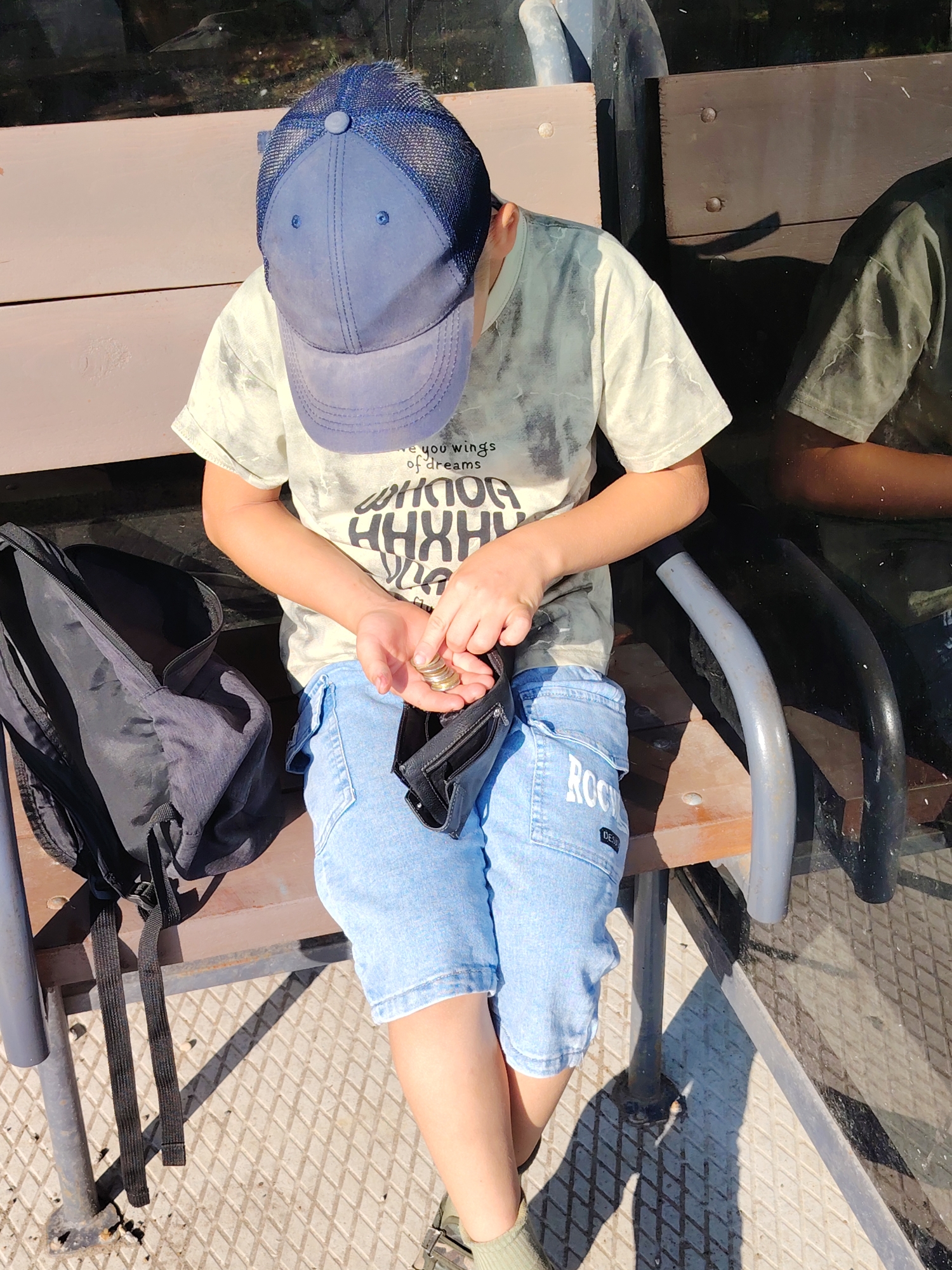 A young person wearing a blue baseball cap and a light-colored t-shirt with a graphic print sits on a wooden bench next to a black backpack. The person is holding coins in their left hand and appears to be counting them. They are also holding a black wallet in their right hand. The bench is situated in an outdoor public space, likely a park or transit stop, with a diamond-patterned metal surface on the ground. The background shows reflections in a window or glass surface, with hints of greenery and urban structures. The overall lighting suggests it is daytime.