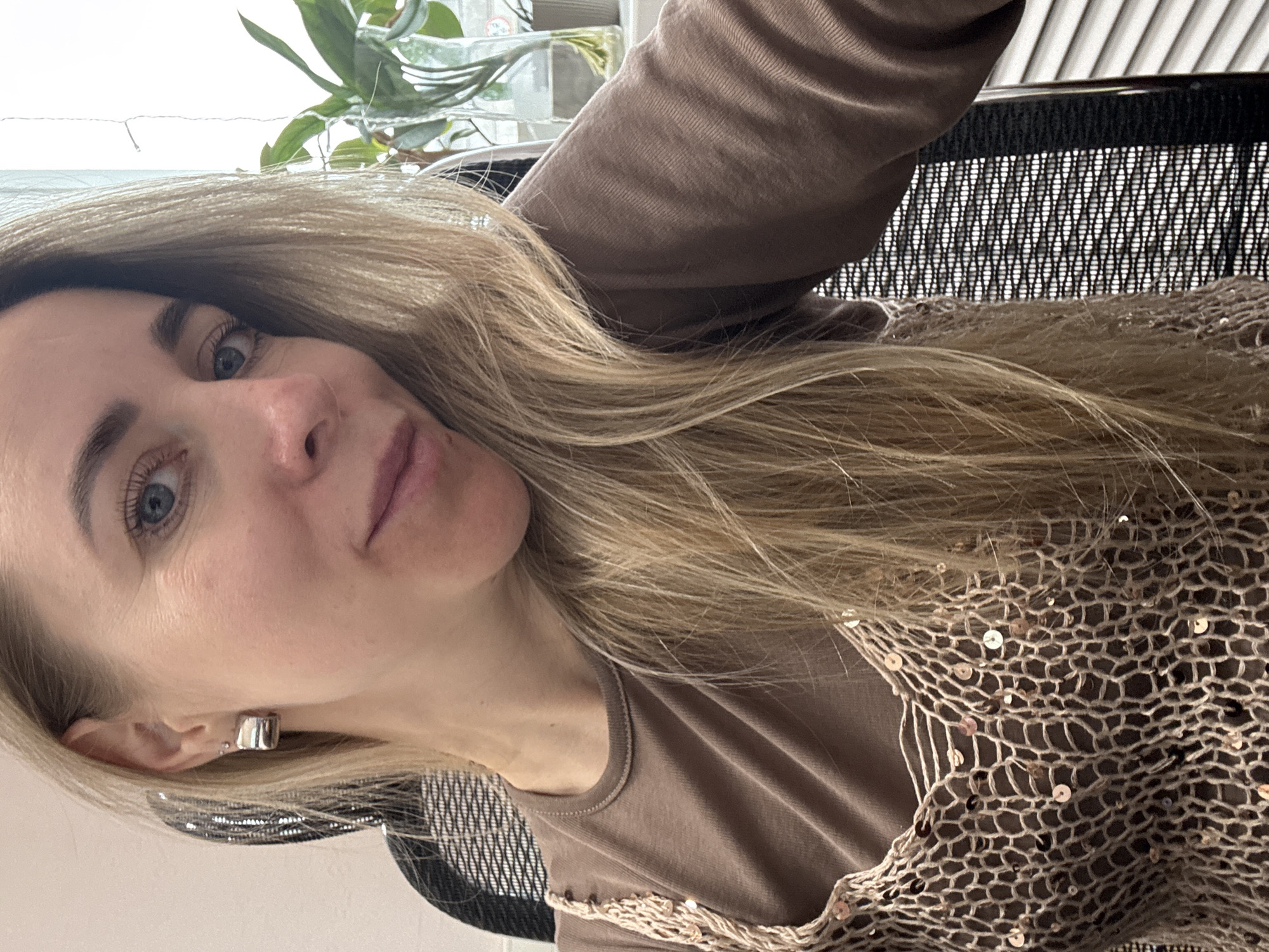A fair-skinned woman with long blonde hair and blue eyes is posing for a selfie. She's wearing a brown t-shirt with a sequined net-like garment over it, and a silver hoop earring. The background shows a partially visible window with a plant on the windowsill and a black mesh office chair. The location is indeterminate, and no evidence suggests the city of Chaikovsky, Russia.