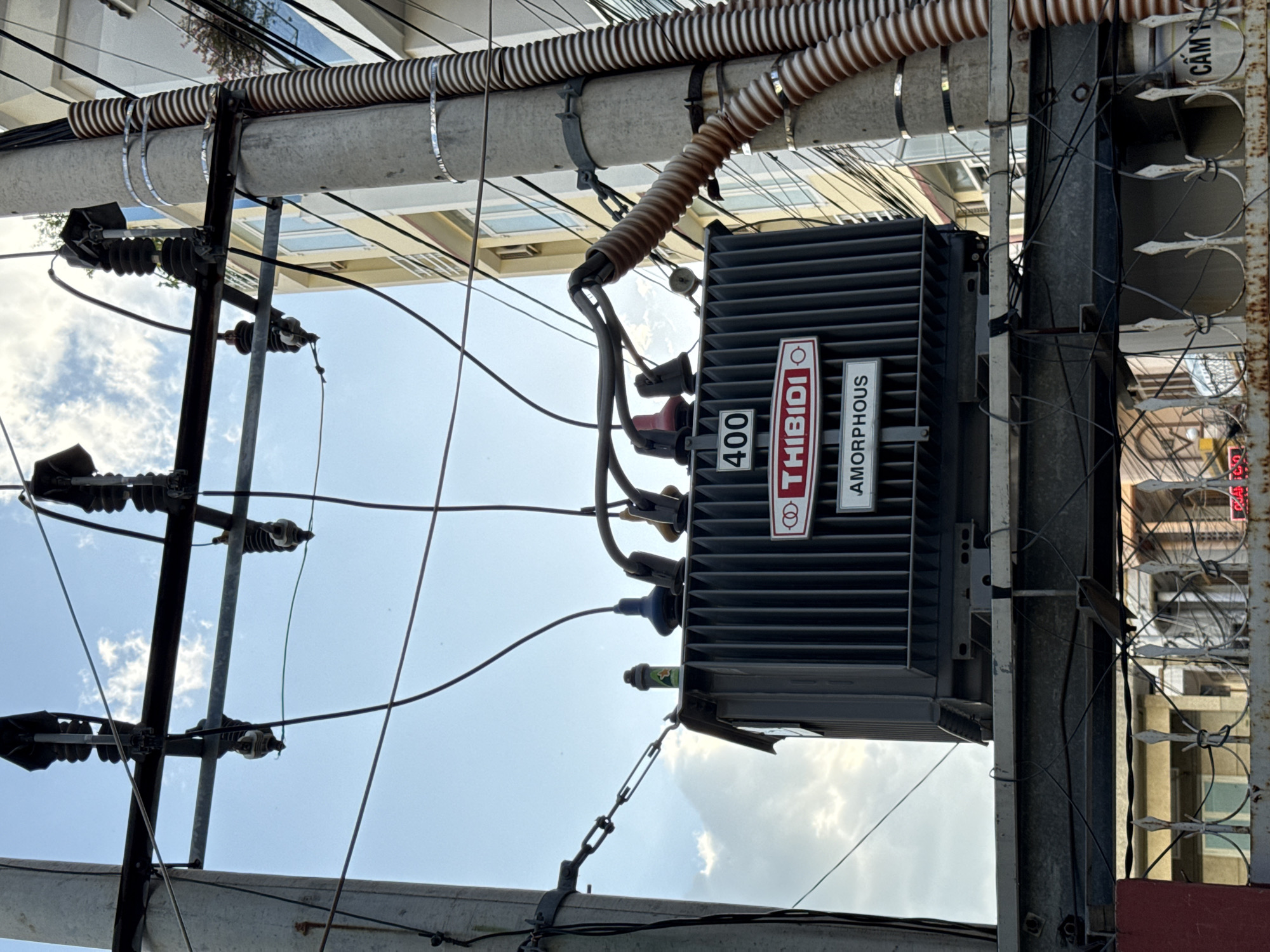 This is a photograph of an outdoor electrical transformer unit mounted on a concrete utility pole. The transformer is dark gray with visible vertical fins and has several labels on its front. One label displays the number "400" in a white rectangle. Below that, a red and white label reads "THIBIDI" with a circular logo. Another label beneath that says "AMORPHOUS". The transformer is connected to overhead electrical wires by thick, insulated cables. The scene is set against a bright blue sky with scattered white clouds. In the background, a light-colored building with multiple windows is visible. The utility pole is also wrapped with corrugated piping. The photograph appears to have been taken in an urban setting, likely a street in Nha Trang, Vietnam, given the presence of typical Vietnamese urban infrastructure. There are no people visible in the image.