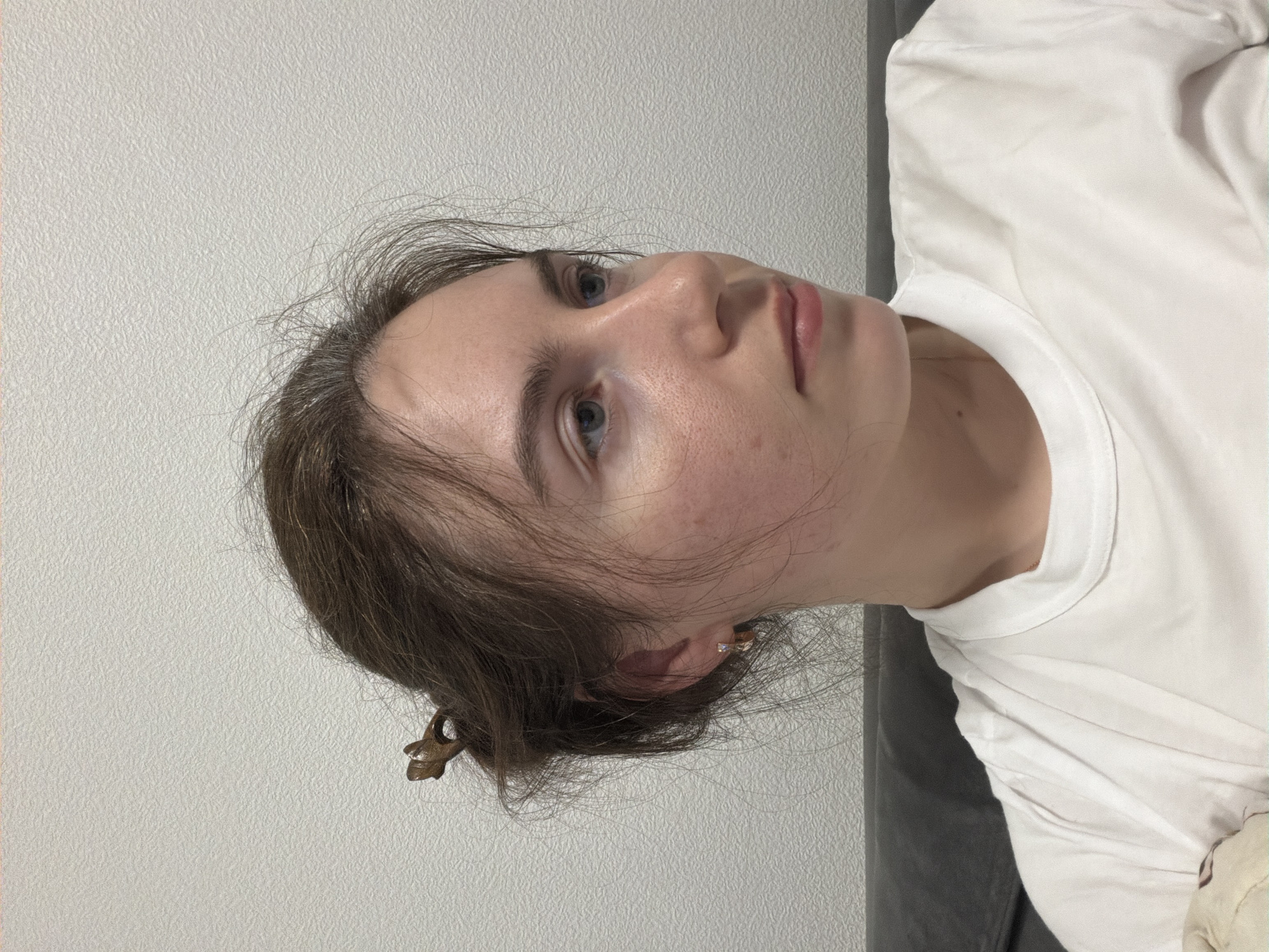 A woman with fair skin, blue eyes, and brown hair pulled back is positioned against a light grey wall. She is wearing a white t-shirt. A small, decorative hair clip is visible in her hair. Her gaze is directed upwards and slightly to the right. The background appears to be a plain wall, suggesting an indoor setting. The image does not provide sufficient information to determine the specific location in Vsevolozhsk, Russia, as there are no identifiable landmarks or contextual clues to establish this.