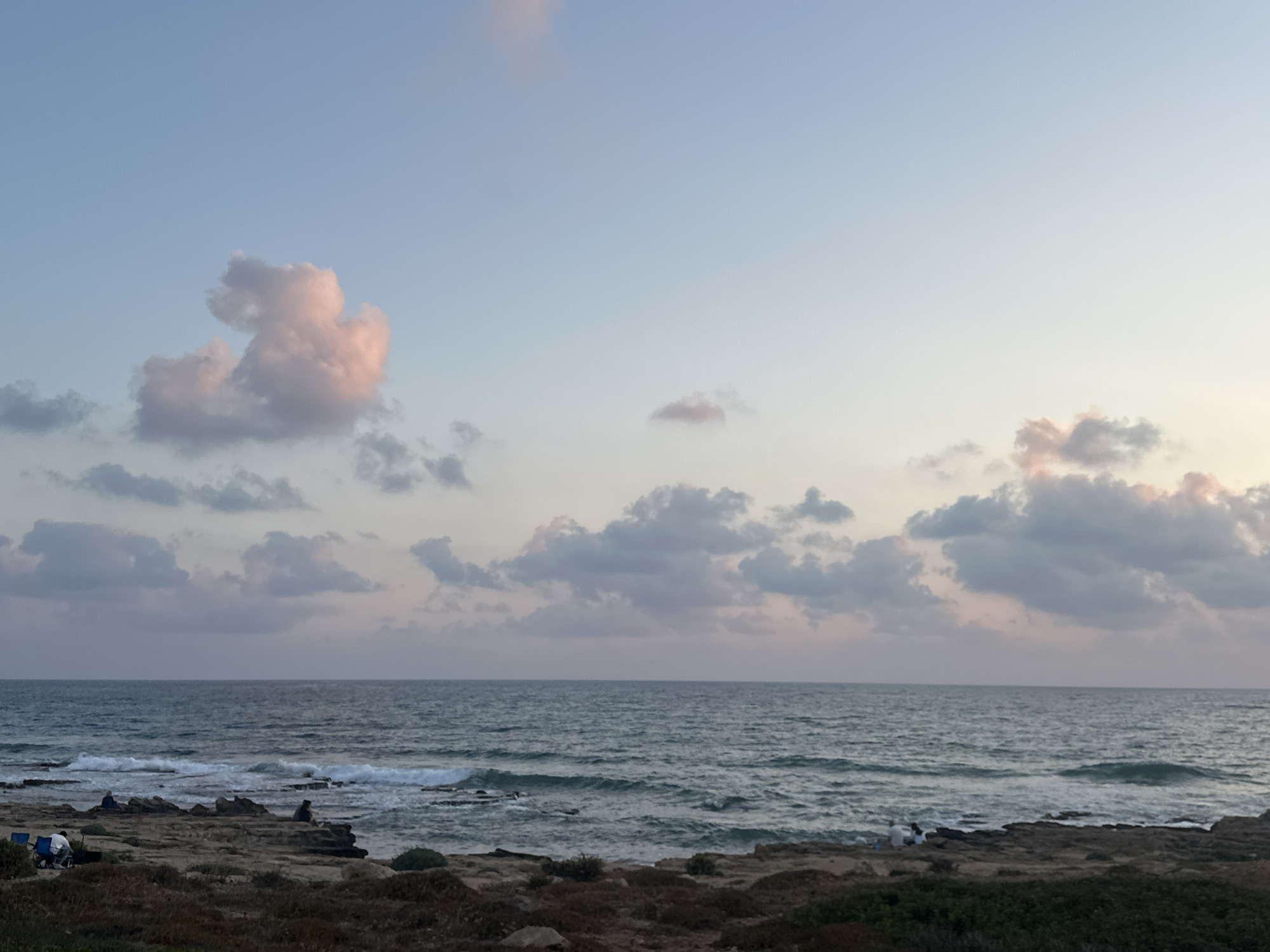 The image depicts a rocky shoreline meeting the Mediterranean Sea, likely in the Mate Asher Regional Council of Israel. The sky is a gradient of soft blues and pinks, indicating either sunrise or sunset, with scattered clouds tinged with the same hues. Several people are visible on the rocks, some sitting in chairs, others appearing to be observing the sea. The water is calm with gentle waves lapping against the shore. The foreground consists of dry brush and scattered rocks.