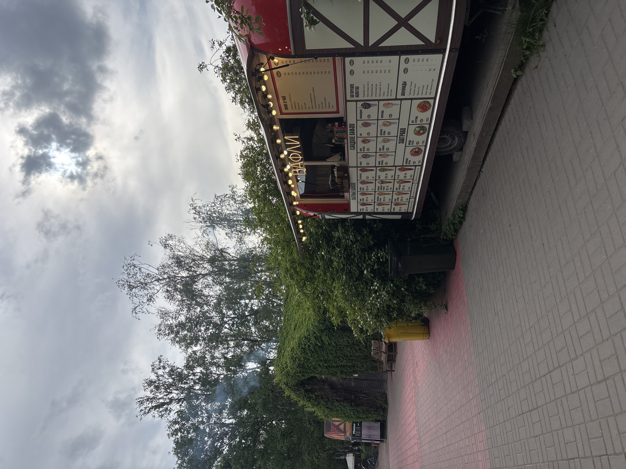 An outdoor food truck, distinctive with its red and white timber-frame-style facade, stands in a pedestrian area, likely a park or recreational zone, in Saint Petersburg, Russia. The scene is set under a cloudy sky, with the sun partially visible, suggesting it's daytime, possibly late afternoon, as string lights on the food truck are illuminated, casting a warm glow.
The main subject is the food truck, which features multiple menu boards detailing its offerings. A prominent neon sign on the side spells out "ВАФЛИ" (Waffles). The menu categories, written in Russian, include "СЫТНЫЕ ВАФЛИ" (Savory Waffles), "СЛАДКИЕ ВАФЛИ" (Sweet Waffles), "КОФЕ И ЧАЙ" (Coffee and Tea), "АВТОРСКИЕ НАПИТКИ" (Signature Drinks), "ЗАВТРАКИ" (Breakfasts), and "ЛИМОНАДЫ" (Lemonades). Specific items like "Кедровый латте" (Cedar latte), "Лавандовый раф" (Lavender Raf), "Грейпфрут бузина" (Grapefruit elderberry), and "Морс клюква" (Cranberry Mors) are listed with prices ranging from 150 to 350 (likely rubles).
The food truck is situated on grey interlocking pavers. Lush green trees and dense vines covering a nearby structure frame the setting, adding to a natural, relaxed atmosphere. A yellow bin is visible near the truck, and benches are partially seen in the background, suggesting a seating area for patrons. Wisps of smoke rise from behind the dense foliage in the distance, indicating some outdoor activity or cooking. No people are visible, but the setup implies an active food service operation.