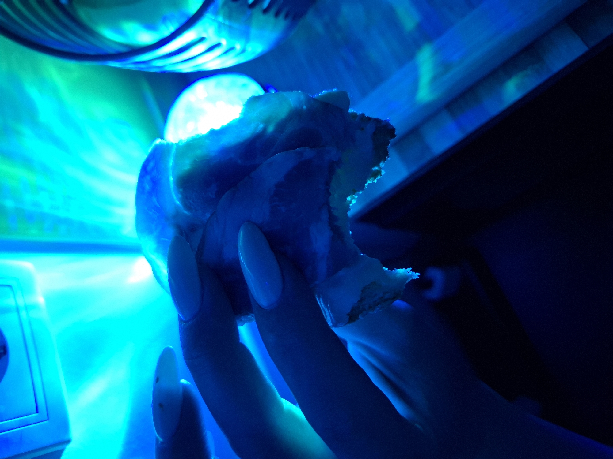 The image captures an indoor scene within the city of Zemun Urban Municipality, Serbia, illuminated predominantly by a vibrant, artificial cyan-blue light. A human hand, featuring manicured long fingernails painted a light color that glows under the blue light, is centrally positioned. The hand holds a partially consumed, irregularly shaped object, consistent with a food item. The object displays a prominent light-colored, possibly white, layer that emits a strong blue luminescence, with a darker, potentially pink or beige layer visible beneath it, suggesting a layered snack or pastry. Visible damage to the object indicates it has been bitten or torn. In the upper left background, a white electrical outlet or control panel is faintly visible on a wall. To the right, a vertically ribbed, reflective object, likely a glass or decorative fixture, catches and reflects the blue light. A dark, flat surface, possibly a table, is present in the lower portion of the frame.