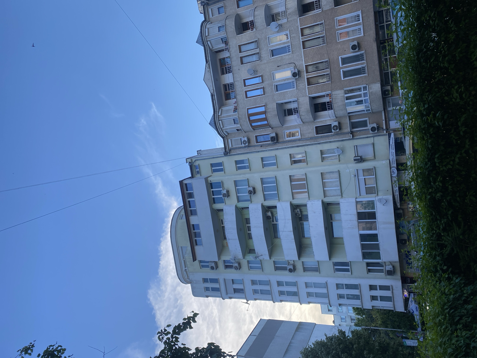 The image shows a street view in Varna, Bulgaria, featuring two apartment buildings against a bright blue sky with scattered clouds. The building on the left is a pale green, multi-story structure with a rounded corner on the top floors. It has numerous windows and balconies, many equipped with air conditioning units. Satellite dishes are visible mounted on the balconies.
To the right of the green building is a light brown, also multi-story apartment building. This building has a more ornate facade with bay windows and decorative cornices. It also features many windows and balconies with air conditioning units and satellite dishes.
The ground floors of both buildings appear to house shops and businesses, indicated by signage visible through the foliage in the foreground. Lush green bushes and trees line the street in front of the buildings. Overhead, power lines crisscross the sky, and a small bird is visible flying in the upper right quadrant. The perspective is from ground level, looking up at the buildings and the sky. The overall impression is an urban residential and commercial area.