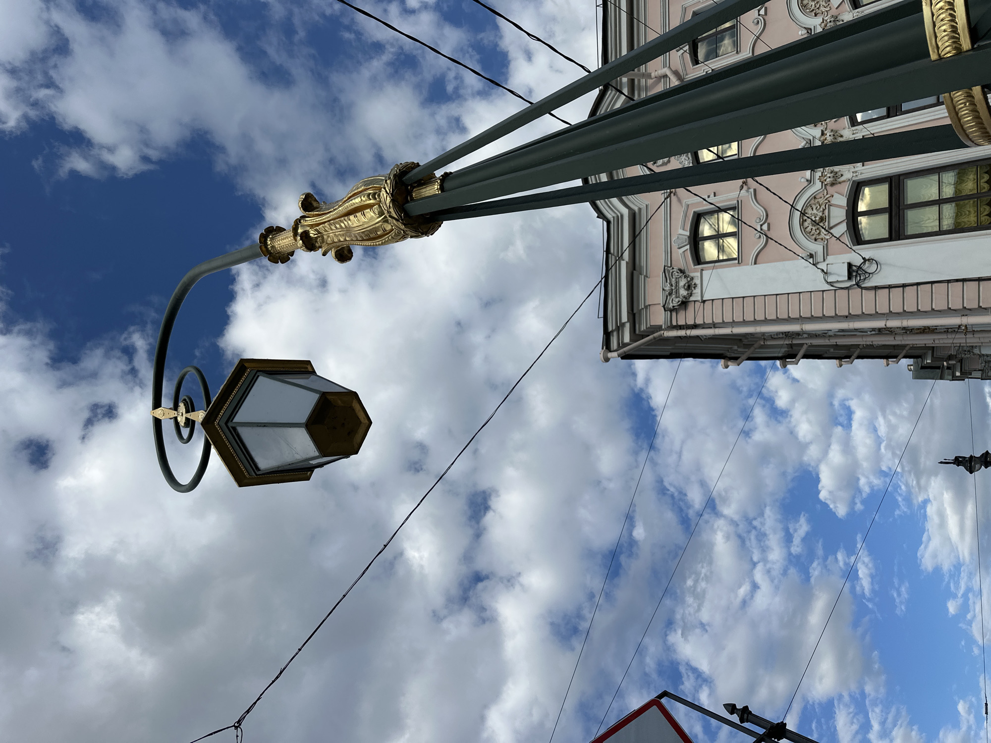 An ornate lamppost with a hexagonal lantern is prominently featured against a blue sky with scattered white clouds. The lamppost is dark green with a decorative gold element near the top. Below the lamppost, a portion of a pale pink building with white trim and arched windows is visible. The building exhibits classical architectural details. Several dark wires are strung across the sky, some appearing to be utility lines. A red and white traffic sign is partially visible on the left side of the frame. The scene appears to be an outdoor urban environment in Saint Petersburg, Russia, likely on a street or bridge, given the presence of the lamppost and overhead wires.