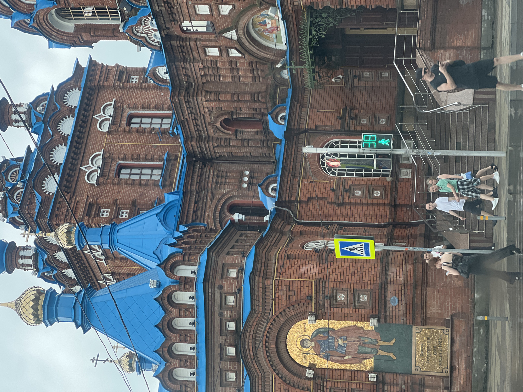 The image depicts the Alexander Nevsky Cathedral in Krasnodar, Russia. The cathedral, made of red brick, features numerous blue domes with gold accents and intricate architectural details. In front of the cathedral, a pedestrian crossing is visible with a traffic light displaying a green walking figure. Several people are crossing the street and walking on the sidewalk. One man is looking at his phone while walking. The sky is overcast, suggesting a cloudy day. The scene appears to be a typical urban streetscape with a prominent religious building.