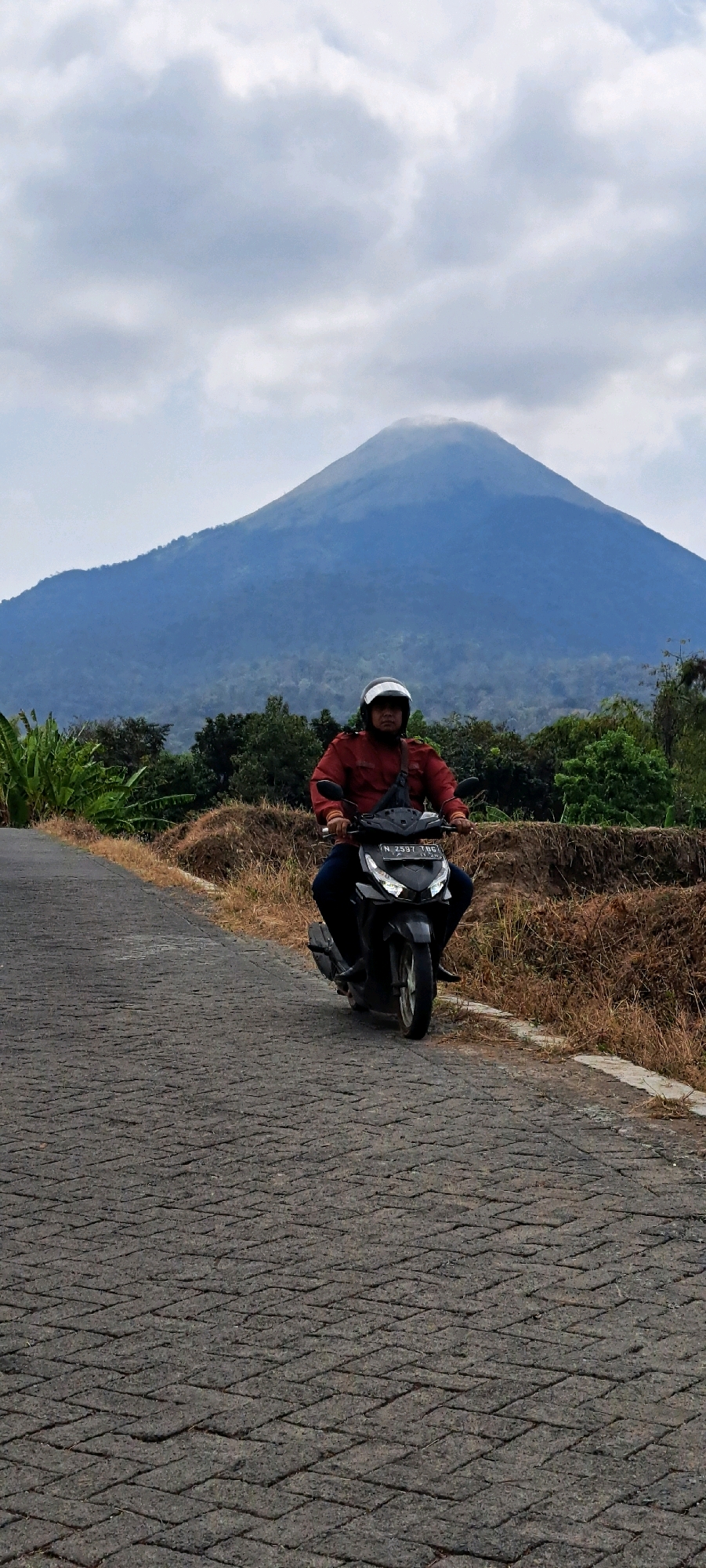 A person wearing a red jacket and white helmet rides a black motorcycle on a cobblestone road in Sumber Suko, Indonesia. The motorcycle has a license plate that reads "N 2597 TBC". In the background, a large, misty mountain looms under a cloudy sky. Lush green trees and dry brush flank the road, indicating a rural setting. The scene captures a moment of travel through a natural landscape.