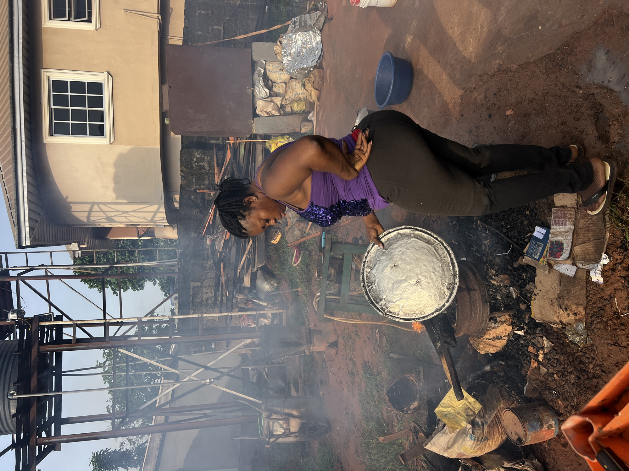 A woman is standing outdoors in what appears to be a backyard or open area, possibly in Nkpor, Nigeria, based on the context provided. She is wearing a purple sequined tank top and dark pants. The woman is holding a large, round, metallic pot or lid, which appears to contain a white, dough-like substance. Her right hand is on her lower back, and she is looking down at the pot.
The environment is somewhat cluttered and industrial. In the background, there is a large metal water tank on a metal stand, along with various pieces of scrap metal and wood. Smoke is visible in the air, suggesting some form of cooking or burning activity. There are also several bags, possibly containing cement or other materials, stacked against a wall. Other objects in the scene include a wheelbarrow, various containers, and debris scattered on the ground, which is a mix of red earth and some patches of mud. The overall impression is of an active, outdoor workspace, likely used for domestic or small-scale industrial activities. The lighting suggests it is either early morning or late afternoon.