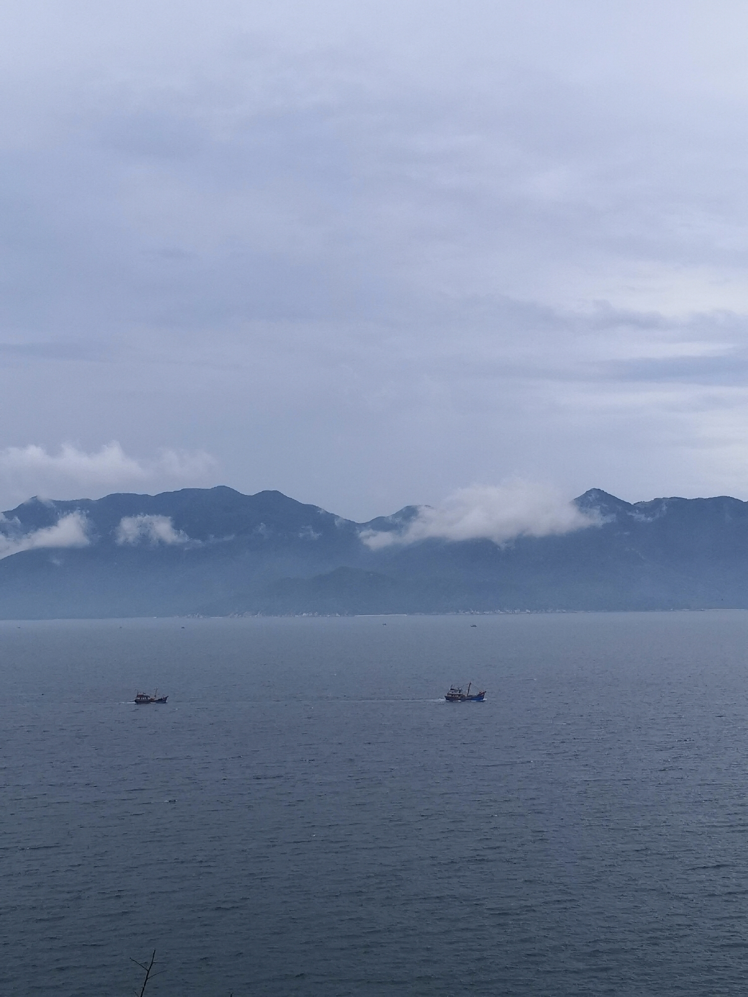 The image shows a seascape with mountains in the background partially obscured by clouds. Two fishing boats are visible on the water. The location is likely in or near Bac Nha Trang Ward, Vietnam, given the mountainous coastal landscape and presence of fishing boats. The sky is overcast.