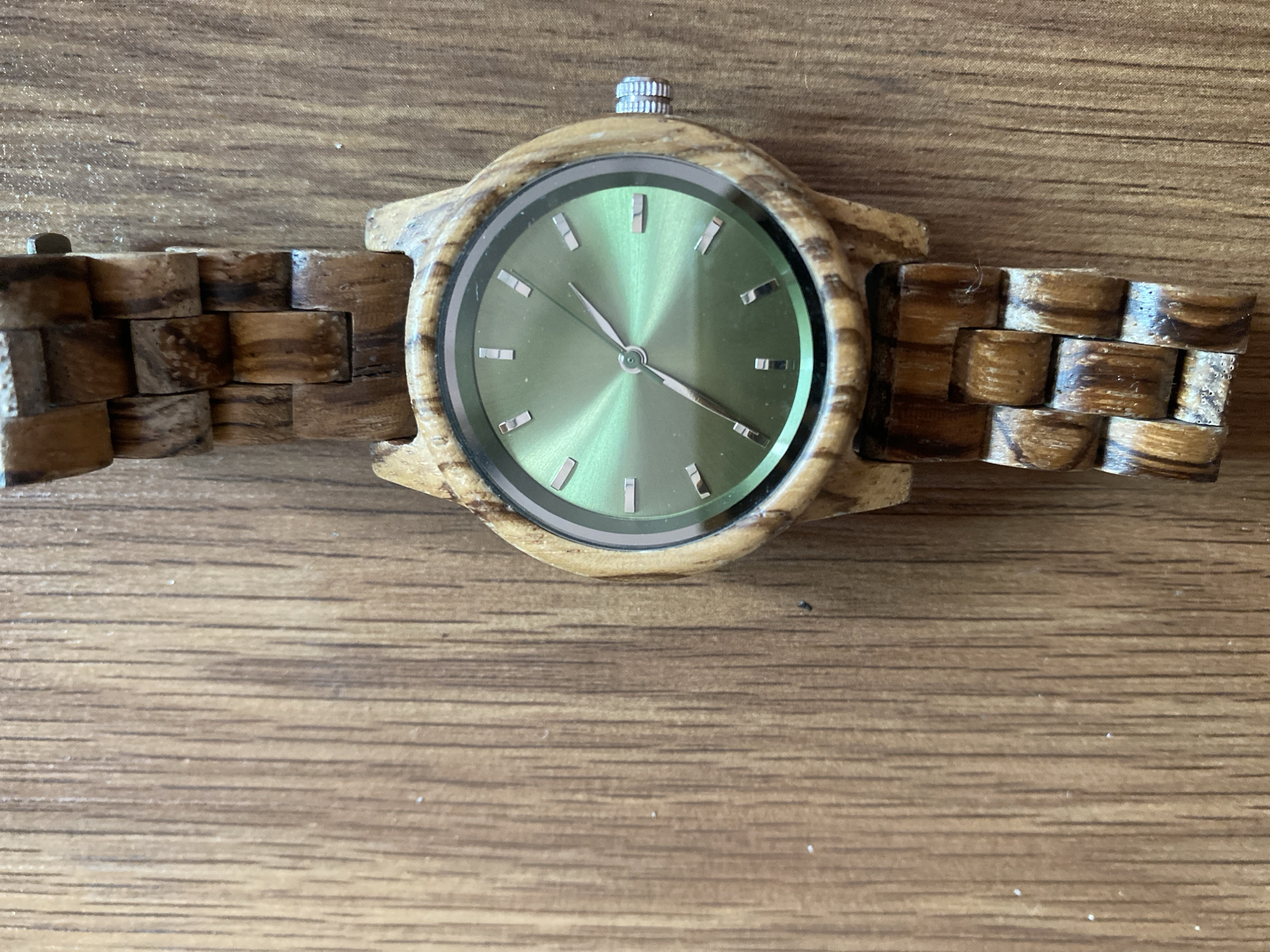 The image shows a wooden watch with a green face lying on a wooden surface. The watch has a woven wooden band. The location is not discernible from the image.