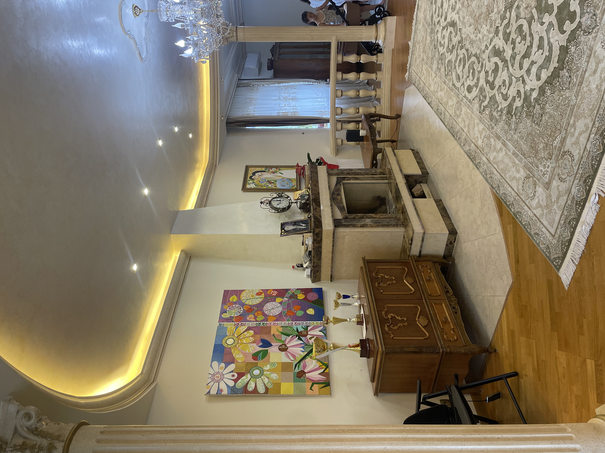 The image shows an opulent living room interior, likely in Almaty, Kazakhstan, judging by the architecture and decor. A large, ornate fireplace dominates the center of the room, made of marble with a decorative mantel. Above the fireplace, a collection of framed artwork and a vintage clock are displayed. To the left of the fireplace, a wooden dresser with intricate carvings holds several trophies. A colorful abstract painting hangs on the wall above the dresser, featuring floral motifs. The room features high ceilings with decorative molding and recessed lighting, as well as a grand crystal chandelier. A large Persian rug with an intricate pattern covers a portion of the wooden floor. In the background, a railing separates the living area from another part of the house, where two young girls are visible, one pushing a stroller. The overall impression is one of wealth and a taste for classical and artistic decor.