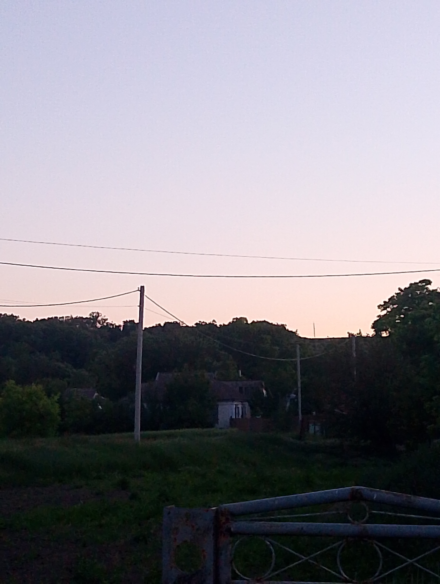 The image depicts an outdoor scene in Ustyvytsia, Ukraine, at what appears to be either dawn or dusk due to the soft, gradient lighting in the sky. The sky is a pale gradient of light blue and pink. In the foreground, a dark, grassy area leads up to a rusted metal gate with circular and crisscross decorative elements. Behind the gate, a sloping hill is covered with dense green trees and shrubs. Several houses with dark roofs are visible nestled among the trees on the hillside. Two utility poles stand upright on the slope, with wires stretched between them and across the frame. The overall impression is a quiet, rural landscape.