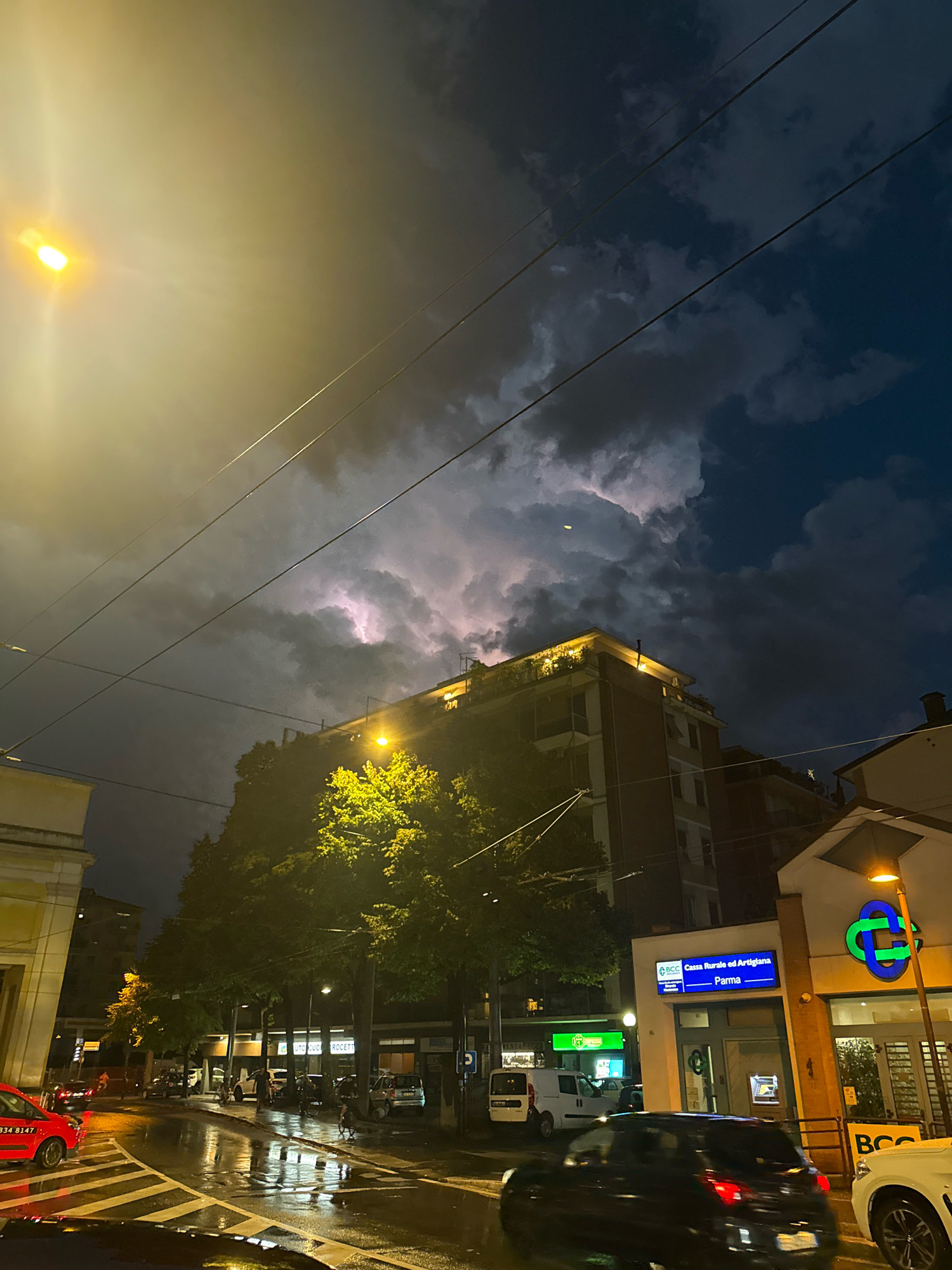 The image captures a street scene at dusk in Parma, Italy, under a dramatic sky illuminated by lightning. A street lamp on the left casts a warm, hazy glow, while the sky above is a mix of dark, stormy clouds and streaks of purple and white lightning. Electrical wires crisscross the frame.
In the mid-ground, a multi-story building with lit windows stands behind a line of trees. The trees, illuminated by streetlights from below, are a vibrant green and cast reflections on the wet street. The street itself is slick with rain, with visible lane markings and reflections of streetlights and buildings.
Several vehicles are present on the road, including a red car on the left, a dark-colored car in the foreground, and a white vehicle on the far right. Other cars and what appear to be motorcycles are parked or moving further down the street.
On the right side of the image, a building with a sign that reads "BCC Cassa Rurale ed Artigiana Parma" is visible. A logo with intertwined green loops is also prominent on this building, along with a streetlamp. The overall atmosphere is one of a city experiencing a thunderstorm, with the lighting and wet conditions creating a dynamic and somewhat moody scene.
