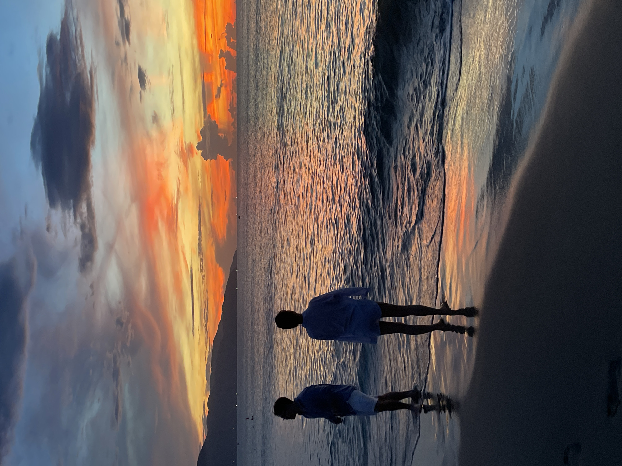 At sunset, two figures stand on the shore of a beach in Da Nang, Vietnam, gazing out at the ocean. The sky is a dramatic canvas of color, with streaks of orange, pink, and yellow painting the clouds against a backdrop of darkening blues. The sun has dipped below the horizon, casting a warm glow over the water and reflecting the vibrant sky onto the wet sand. The figures, silhouetted against the fading light, appear to be a man on the left wearing a blue shirt and white shorts, and a person on the right wearing a loose blue shirt and shorts. They are standing close to the water's edge, with small waves lapping at their feet. In the distance, the silhouette of a mountain range is visible, adding depth to the coastal landscape. The overall scene is one of serene beauty and quiet contemplation, capturing the tranquil end of a day in Da Nang.
