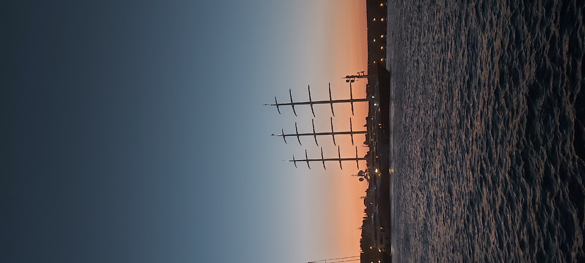 The image depicts a harbor scene at dusk in the Municipal Unit of Rhodes, Greece. Three masts of a large sailing vessel, likely a yacht or schooner, are prominent against a gradient sky transitioning from deep blue at the top to a warm orange-pink near the horizon. Several other yachts are moored in the foreground, their lights illuminated. A lit waterfront or pier with regularly spaced lights is visible in the background, suggesting a developed coastal area. The water in the foreground is choppy with a dark, textured surface reflecting the ambient light. The overall atmosphere is serene and highlights the transition from day to night.