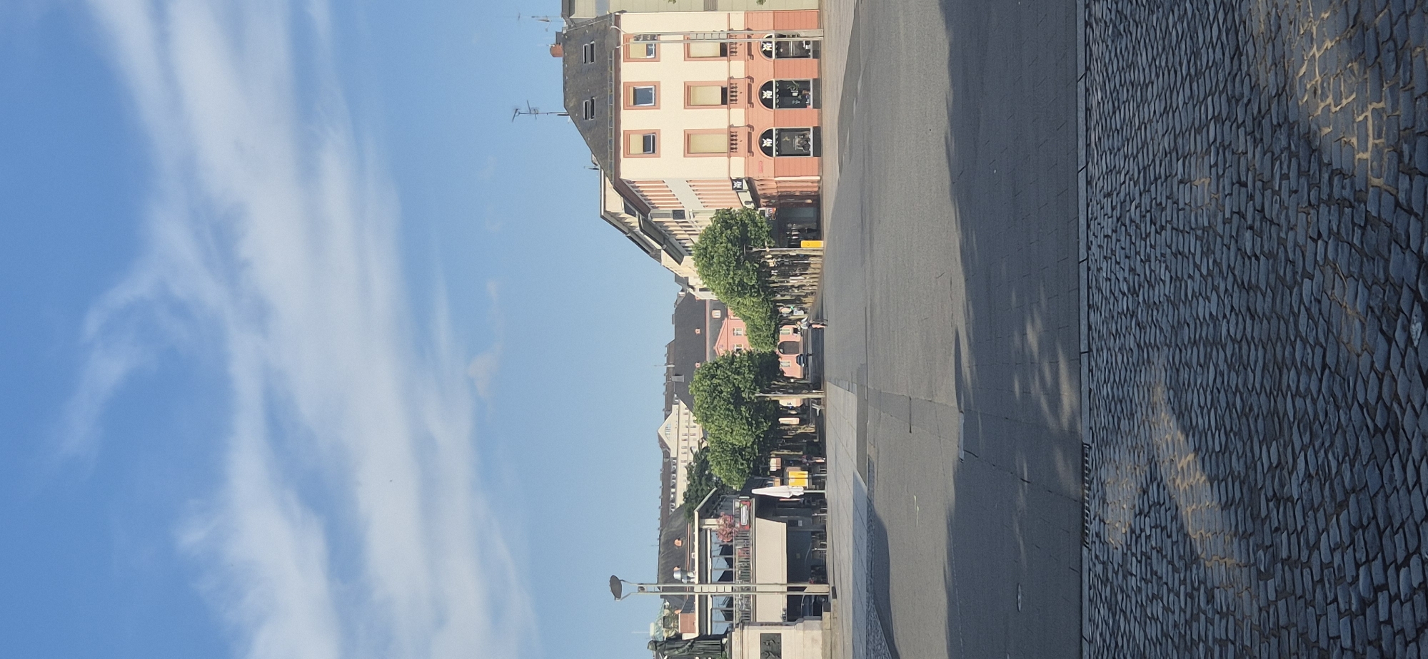 This is a medium-shot, eye-level, vertical, outdoor, daytime photograph taken in the city of Mainz, Germany, likely in a public square. The sky is a clear blue with scattered wispy white clouds. The foreground is paved with dark gray cobblestones, with a visible pattern of light and shadow cast by nearby trees.
The midground is a large, open paved area with a lighter gray surface, transitioning to cobblestones at the very bottom of the frame. A sidewalk lined with evenly spaced, mature trees with lush green foliage runs through the center of the square. On the left side of the square, a dark statue on a pedestal is partially visible, along with a multi-story building featuring a balcony with umbrellas and seating, possibly a cafe or restaurant.
The background consists of a row of buildings. On the right, a pastel pink and cream building with multiple floors has dark sloped roofs and visible windows, some with light-colored awnings. There are antennae on the roof. To the left of this building, a series of other buildings with dark roofs and lighter facades are visible. In the distance, a large building with a classical facade and a central triangular pediment can be seen.
A single person is visible in the midground, riding a bicycle down the tree-lined avenue. There are a few other indistinct figures and objects near the cafe area. The lighting suggests it is morning or late afternoon, with distinct shadows cast across the square. The overall impression is one of a quiet, well-maintained European city square on a bright day.