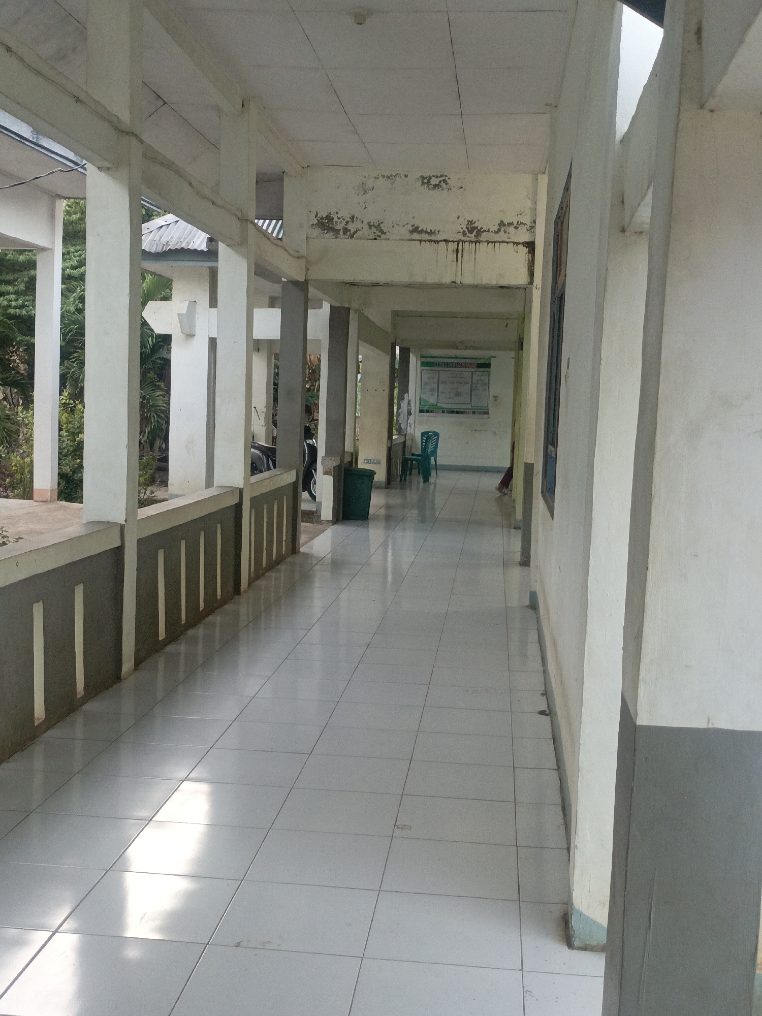 The image depicts an outdoor covered walkway or corridor in Solor Barat, Indonesia. The walkway has a tiled floor, and the walls on the right are concrete. On the left, there is a railing with vertical decorative elements, and beyond that, lush green foliage is visible. Several concrete pillars support the roof of the walkway. In the distance, a green trash can is positioned near a motorcycle. Further down the corridor, a green chair is placed in front of a notice board mounted on the wall. The scene suggests a public building or institution, possibly a school or community center, given the presence of the notice board and the functional, albeit weathered, architecture. The lighting indicates it is daytime, with sunlight casting reflections on the tiled floor.