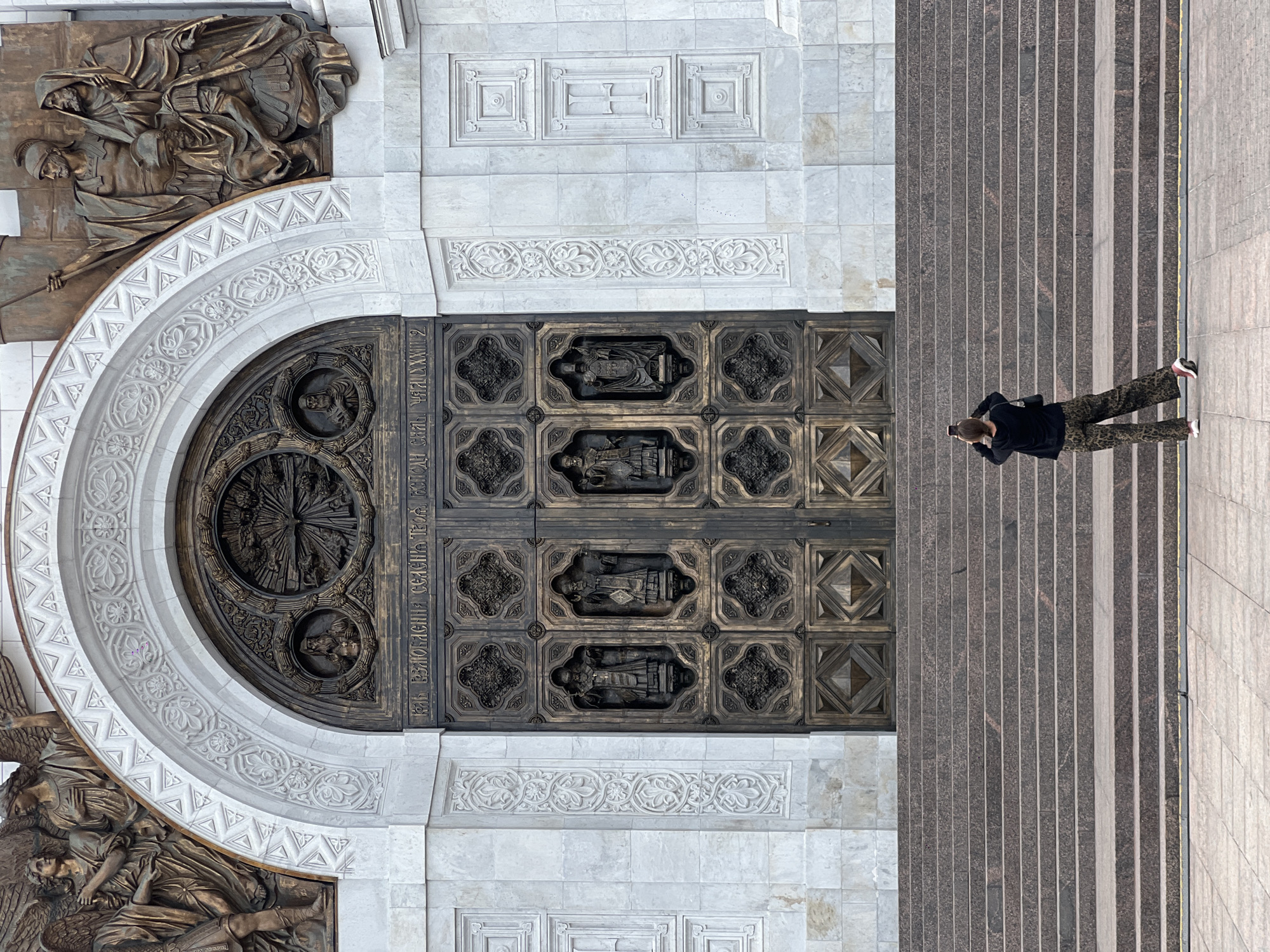 A high-angle, full shot reveals the ornate entrance of the Cathedral of Christ the Savior in Moscow, Russia. A person stands on the adjacent stairs, appearing to photograph the cathedral entrance.
The cathedral entrance features a large, arched, decorative bronze door with intricate carvings and sculpted figures. Adjacent to the door are white stone walls with decorative carvings. To the right of the entrance are a series of steps leading up to the entrance level. These steps are primarily brown and grey with light grey or white stones interspersed.
A person is positioned on the steps, dressed in dark clothing, wearing camouflage-patterned pants and white shoes. They are facing the door and appear to be holding a device (possibly a phone) up to capture a photograph.