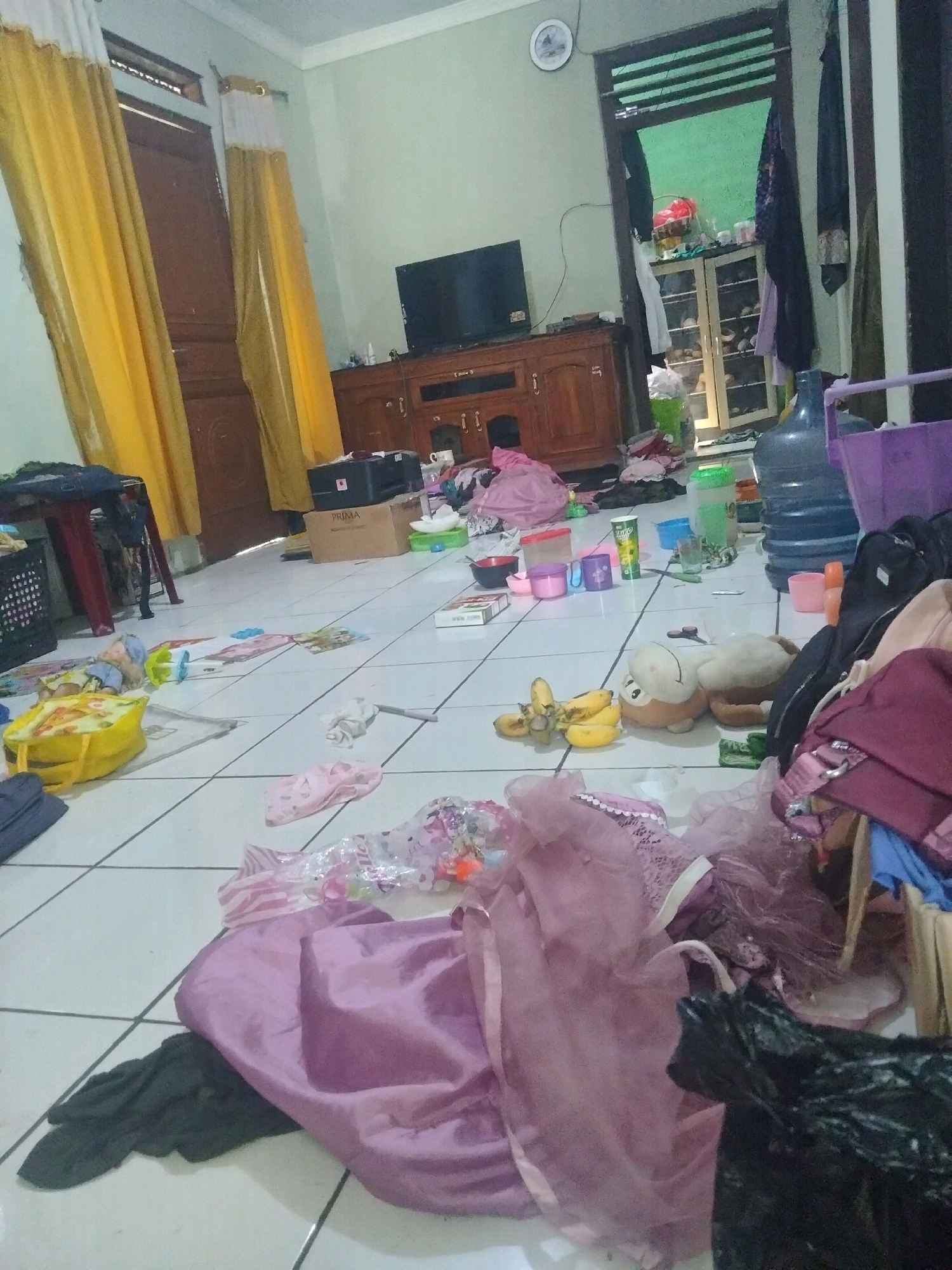 The image depicts a messy room, likely a living area, located in Suka Sari, Indonesia. The floor is covered with scattered clothing, toys, a stuffed animal, bananas, and other miscellaneous items. A large blue water dispenser is visible, along with various plastic containers and bags. A television set is mounted on a wooden cabinet. In the background, a doorway leads to another room with shelves stocked with items. Yellow curtains hang on the left side of the frame. The overall impression is one of disarray and clutter.