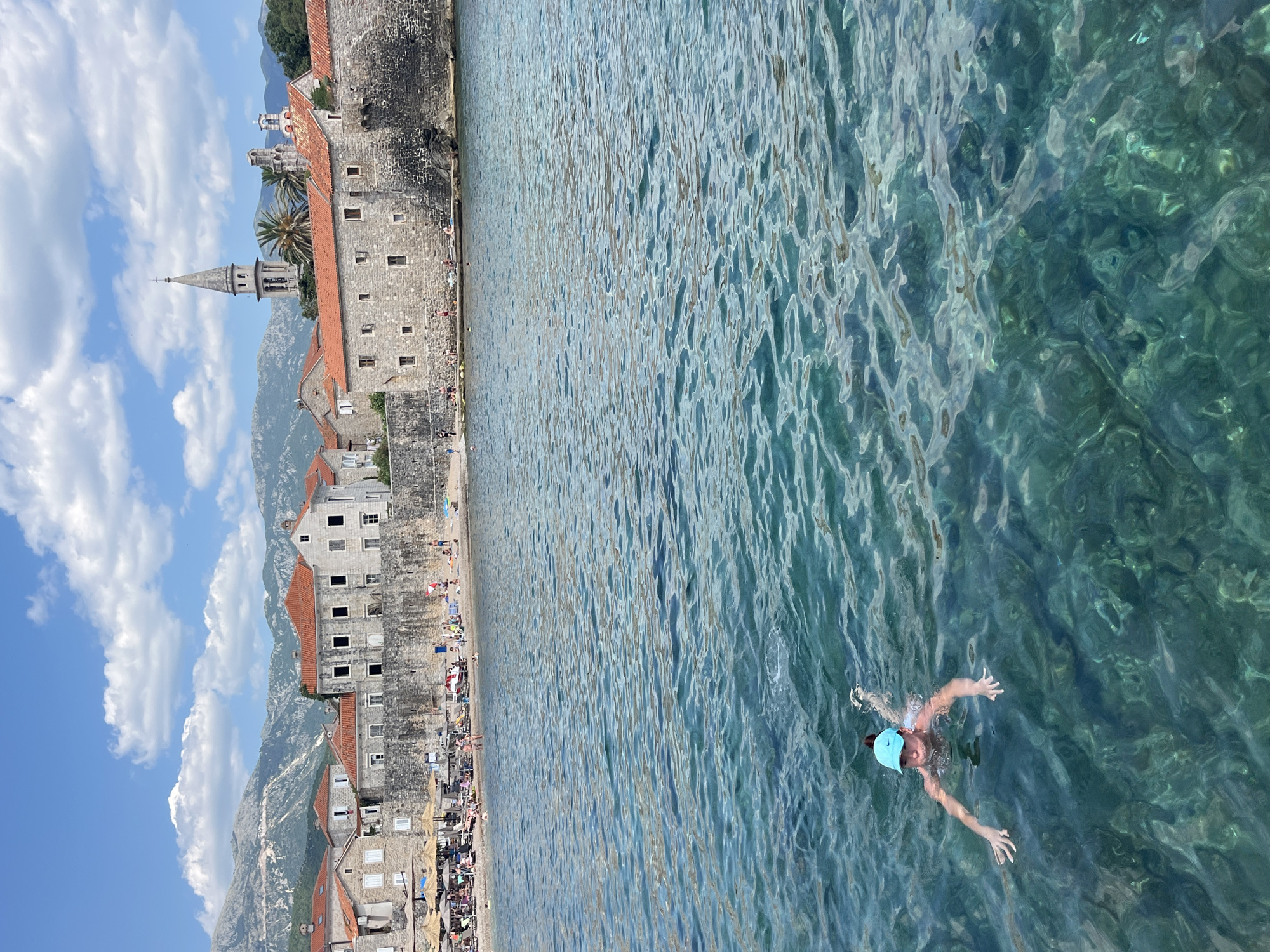 The image depicts a person swimming in the clear, turquoise waters of the Adriatic Sea, with the old town of Budva, Montenegro, as a backdrop. The old town is characterized by its ancient stone buildings with red-tiled roofs, a prominent bell tower, and lush green trees. The foreground is dominated by the rippling water, revealing a sandy seabed beneath. In the mid-ground, the beach is dotted with beach umbrellas and sunbathers enjoying the sunny day. The background features a range of mountains under a blue sky with scattered white clouds.