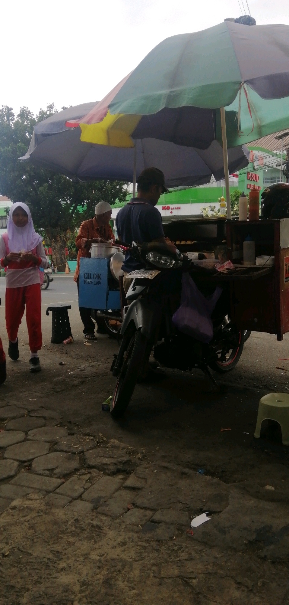 A street food vendor stall is set up under a multi-colored umbrella. A man wearing a dark blue shirt and a baseball cap is preparing food behind the stall, which is mounted on a motorcycle. An older man in an orange shirt and white cap stands to the left of the vendor, next to a blue container labeled "CILOK Masa Lalu". A young girl wearing a light purple hijab and red pants stands to the far left, looking at the viewer and holding something in her hands. The stall is equipped with various containers and bottles, including sauce dispensers. In the background, a building with green signage is visible. The ground is paved with cobblestones.
