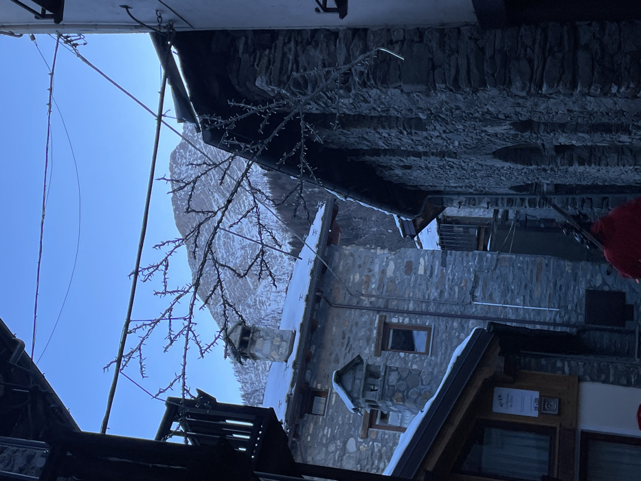The image captures a winter scene in a mountain village, likely Courmayeur, Italy, as indicated by the context. The photograph is oriented vertically (rotated 90 degrees counter-clockwise from its natural state), showing a view upwards through a narrow street or passage.
In the corrected orientation, the scene depicts stone and wooden buildings lining the left and bottom edges of the frame, some with snow accumulated on their dark roofs. One building on the left features a stone chimney and small windows. Overhead, numerous wires and cables crisscross the sky, alongside a decorative structure resembling illuminated branches or string lights, suggesting a festive or winter ambiance.
The background reveals a magnificent snow-covered mountain peak bathed in bright sunlight, which creates a prominent lens flare in the upper-left sky (after correction). The sky itself is a clear, bright blue. A small portion of a vibrant red object, possibly a person's clothing or another decoration, is visible on the far right.
On the wooden facade of one of the buildings, a white sign is visible, displaying "CERTIFICATO DI ECCELLENZA" (Certificate of Excellence) and the "TRIPADVISOR" logo with "2017," along with a "q-cert" logo. Another small sign indicates "Wi-Fi." The image evokes a sense of peaceful, clear winter weather in an Alpine village.