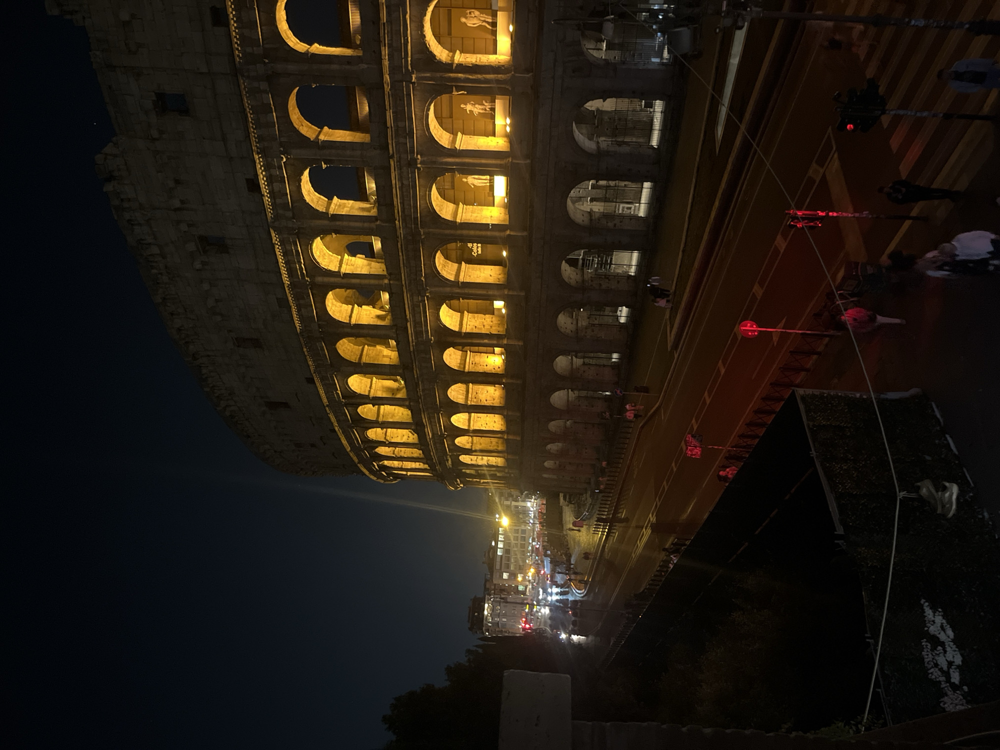 This night-time photograph captures the majestic Colosseum in Rome, Italy, bathed in a warm, artificial glow against a deep blue-black sky. The iconic ancient amphitheater dominates the left portion of the frame, its multi-tiered arcades spectacularly illuminated from within, casting a golden hue that accentuates the rugged texture of its aged stone façade. Within several of the upper arches, faint figures, possibly statues or architectural elements, are visible, adding to the historical richness.
Running parallel to the Colosseum in the mid-ground is a bustling city street, featuring multiple lanes of traffic. Several red traffic lights are clearly visible, indicating a pause in traffic flow and allowing for pedestrian crossing. The road surface reflects some of the scattered artificial lights from street lamps and distant buildings, creating a sense of an active urban environment.
On the right side of the image, a sidewalk or pedestrian crossing is occupied by several indistinct figures, likely pedestrians. Their presence adds a human element to the scene, suggesting people are either visiting the landmark or simply navigating the city streets at night. Further down the street, more distant city lights twinkle, hinting at the expansive urban landscape of Rome. A small sign, partially legible as "Colle...", can be faintly seen within one of the illuminated arches of the Colosseum, possibly indicating an exhibit or information point. The photograph captures a serene yet vibrant evening atmosphere, where ancient history seamlessly coexists with modern city life.