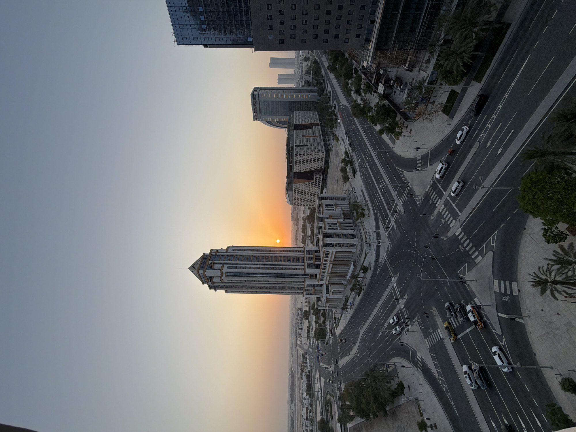 This image shows a cityscape at dusk in Lusail, Qatar. The sky is a gradient of pale orange near the horizon, transitioning to a soft blue. A large, modern skyscraper with a decorative top stands prominently in the mid-ground, partially silhouetted against the sunset. Several other contemporary buildings, including a curved structure and a rectangular building with a patterned facade, are visible in the background. The foreground features a wide, multi-lane intersection with cars navigating through it. Pedestrian crossings are marked with white lines, and traffic lights are visible. The area is lined with trees, including palm trees, and sidewalks. The overall impression is one of an organized, developing urban environment bathed in the warm light of the setting sun.