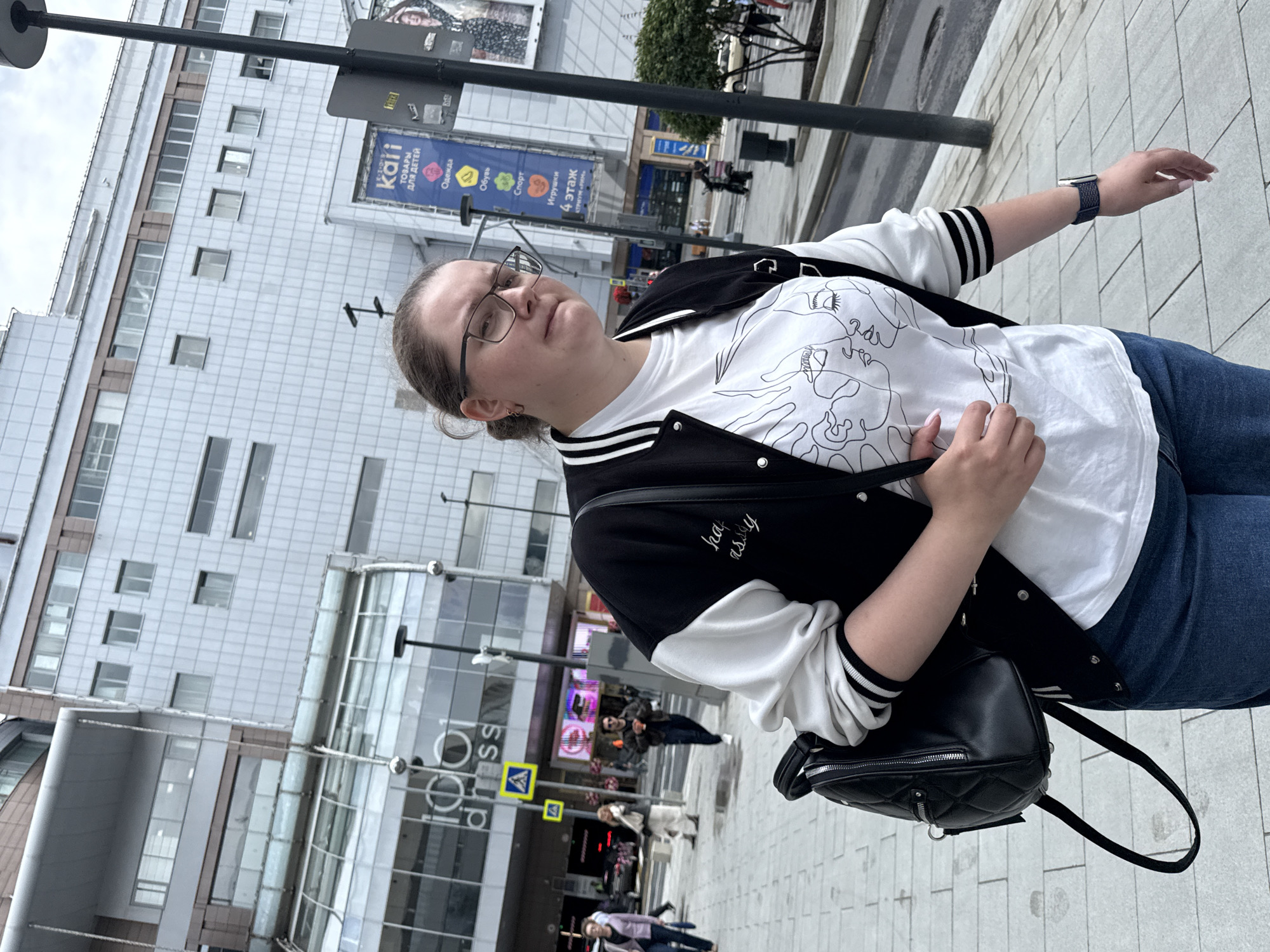 A medium shot captures a woman walking on a paved city sidewalk in Moscow, Russia. She is wearing a white t-shirt with a minimalist line drawing of two faces on it, a black and white bomber jacket, and blue jeans. She carries a black quilted backpack slung over her left shoulder. The woman wears glasses and her hair is pulled back. To her right, a tall black lamppost stands prominently. In the background, a large, modern building with a grey, textured facade is visible. Various signs are affixed to the building, including one in Russian advertising "Kari" children's goods. Other pedestrians and a motorcycle are also present in the background. The sky is overcast.