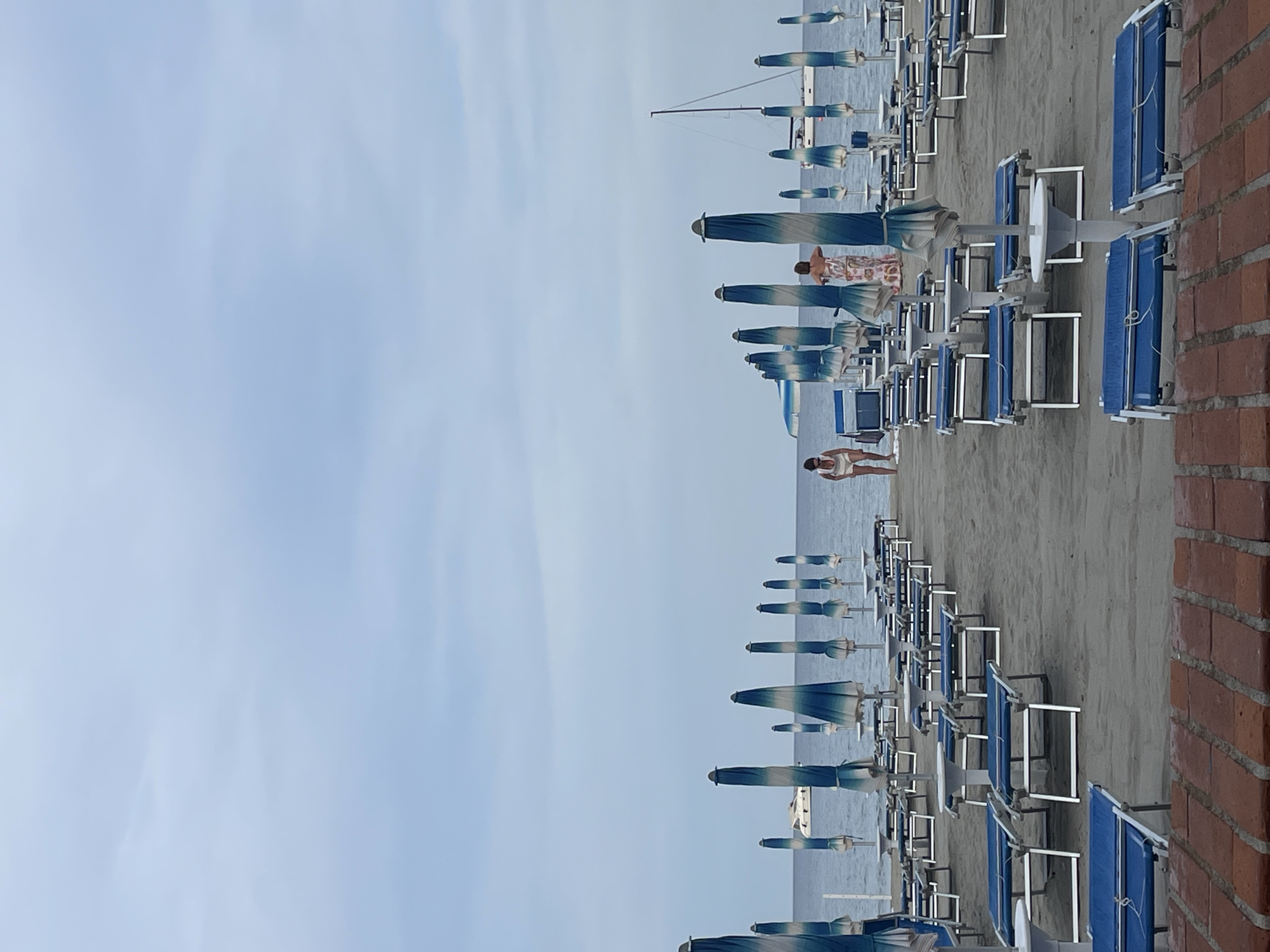 The image captures a beach scene in Laigueglia, Italy. Rows of blue beach chairs and striped umbrellas stretch along the sandy beach, suggesting a resort or private beach area. The sea is visible in the distance, with a sailboat present. A few individuals are seen on the beach, one walking and another reclining on a beach chair. A brick structure borders the beach area. The sky is overcast.