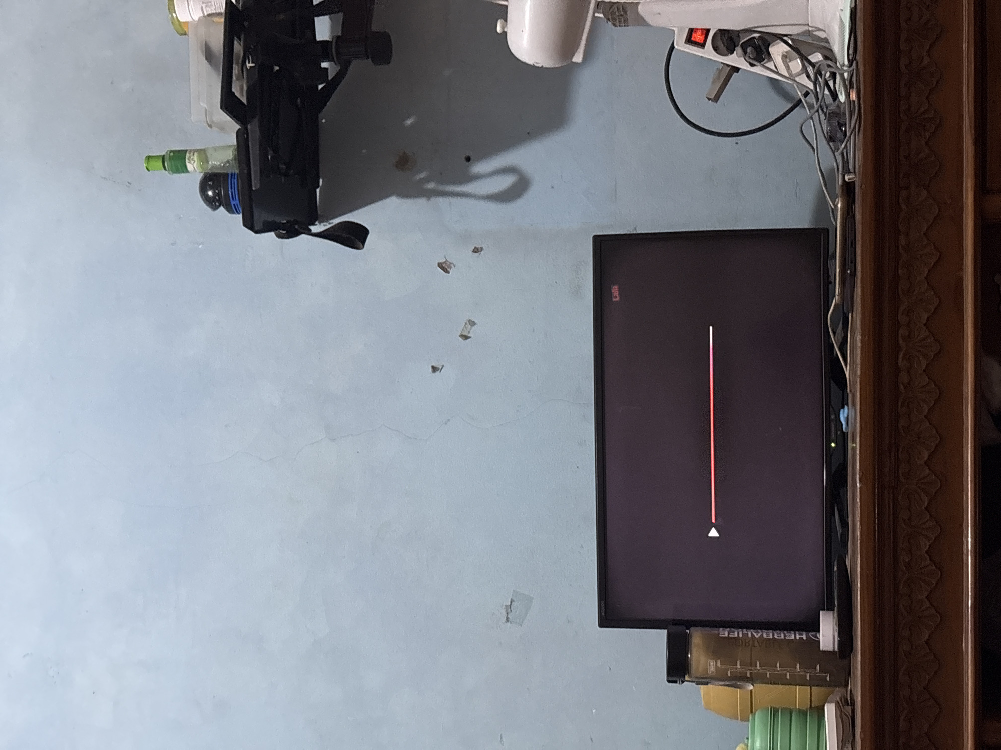 The image shows a dimly lit room with a light blue, textured wall. A television screen in the center displays a loading bar, indicating a video is playing or buffering. On the left side of the television, there are several plastic containers, including a tall, brown bottle with "HERBALIFE" printed on it. To the right of the television, a white electric fan is visible, with various cords and plugs connected to it. A shelf is mounted high on the wall, holding a few items, including a green bottle and a black object. The room appears to be domestic, possibly a living area or bedroom. There are no people visible in the image, and no discernible actions are taking place beyond the loading bar on the screen. The scene is set in Palimanan, Indonesia.